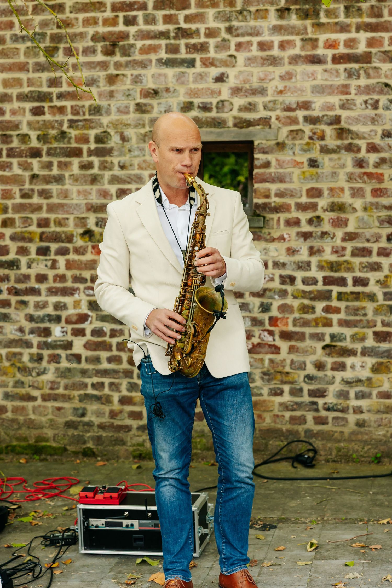 A bald musician in a white blazer plays saxophone against a rustic brick wall at an outdoor event.