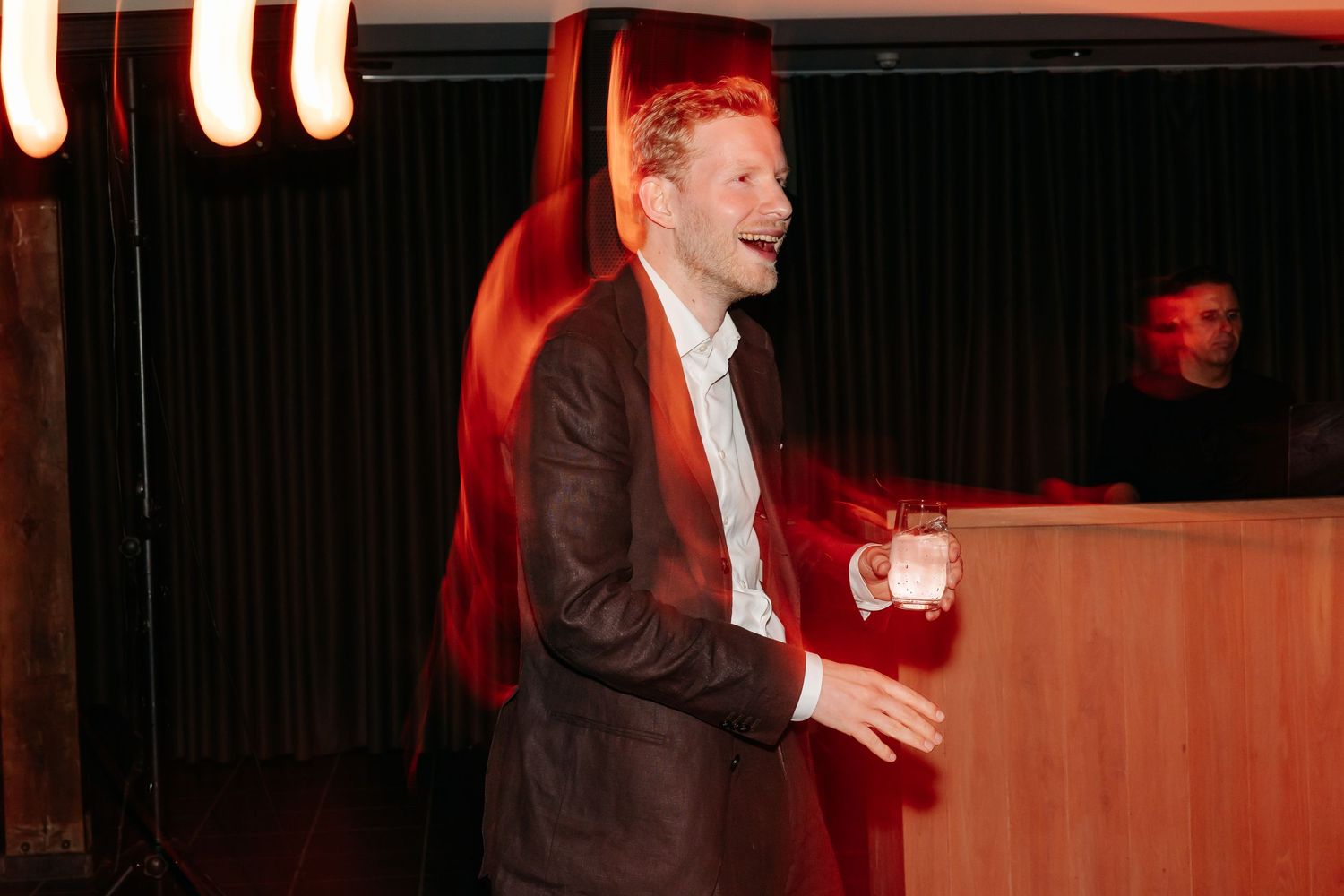 Smiling man in brown suit holding a drink at an evening event with red lighting.