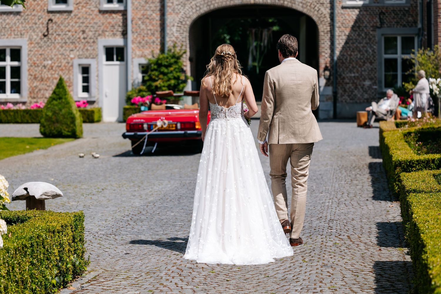 Bride and groom walking hand in hand toward vintage red car at elegant estate venue.