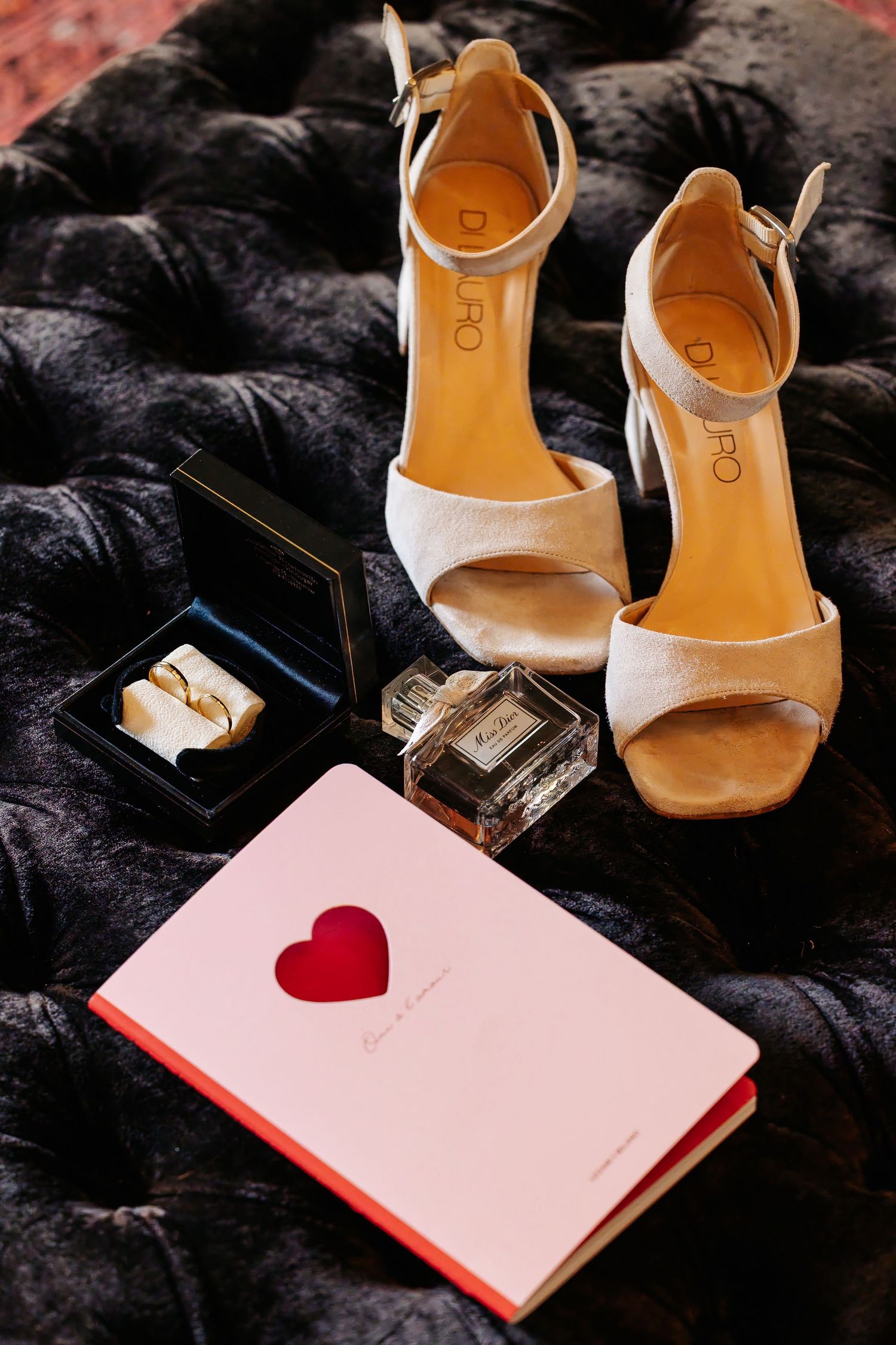 Bridal accessories flat lay with gold heels, wedding rings, perfume, and pink heart notebook on dark velvet.