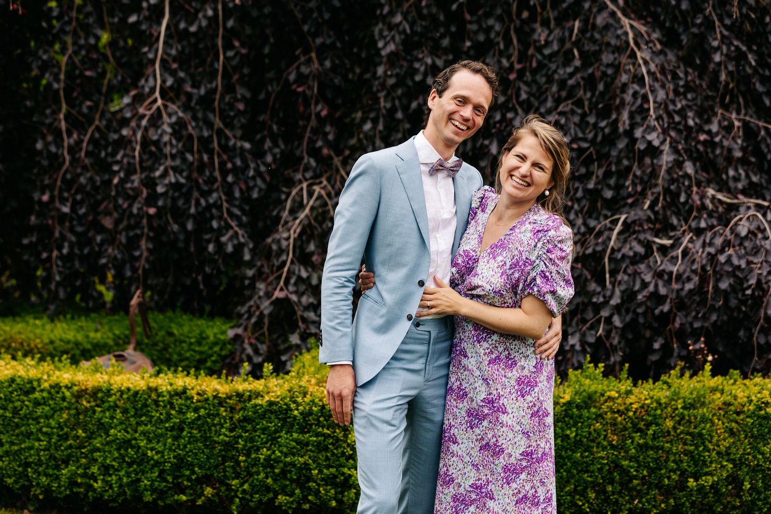 Groom in light blue suit and woman in purple floral dress smiling together in a lush garden setting.