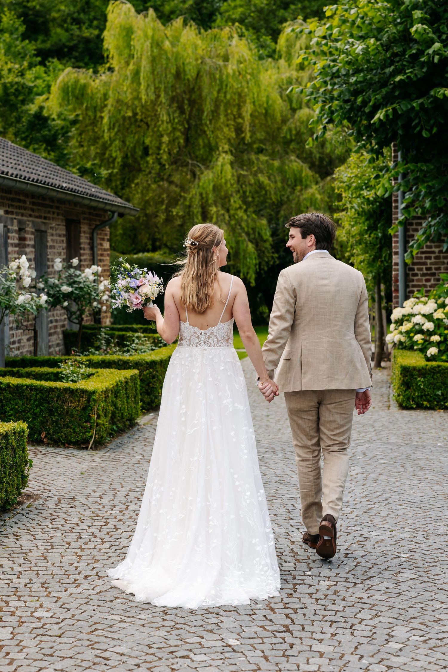 Bride and groom walk hand-in-hand on cobblestone path surrounded by lush greenery at outdoor wedding venue.