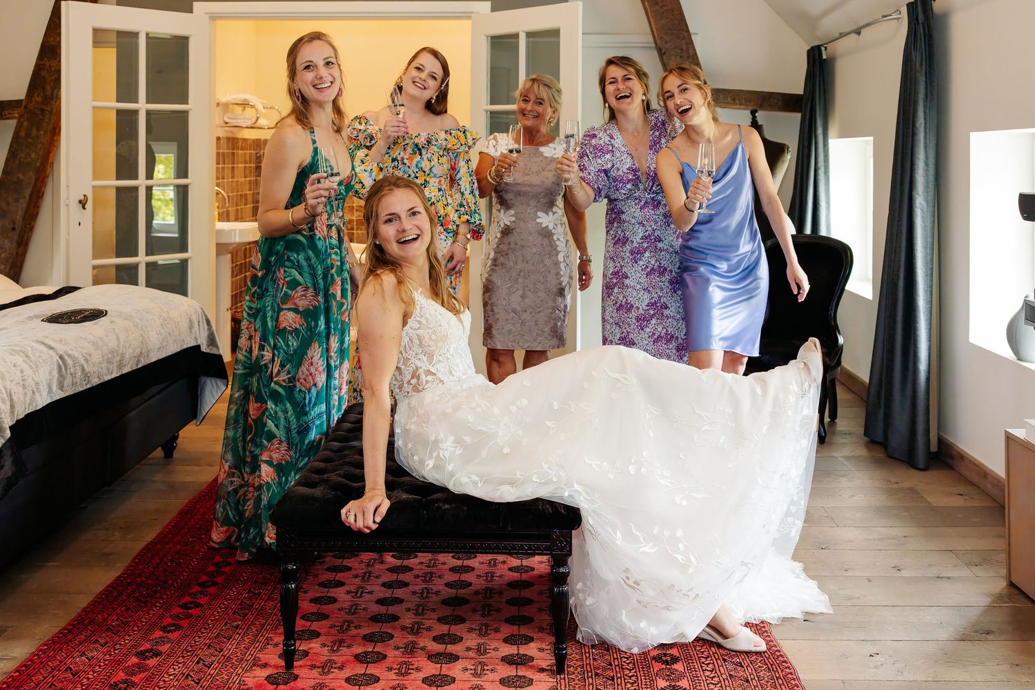 Bride seated with five bridesmaids posing joyfully in rustic bridal suite.
