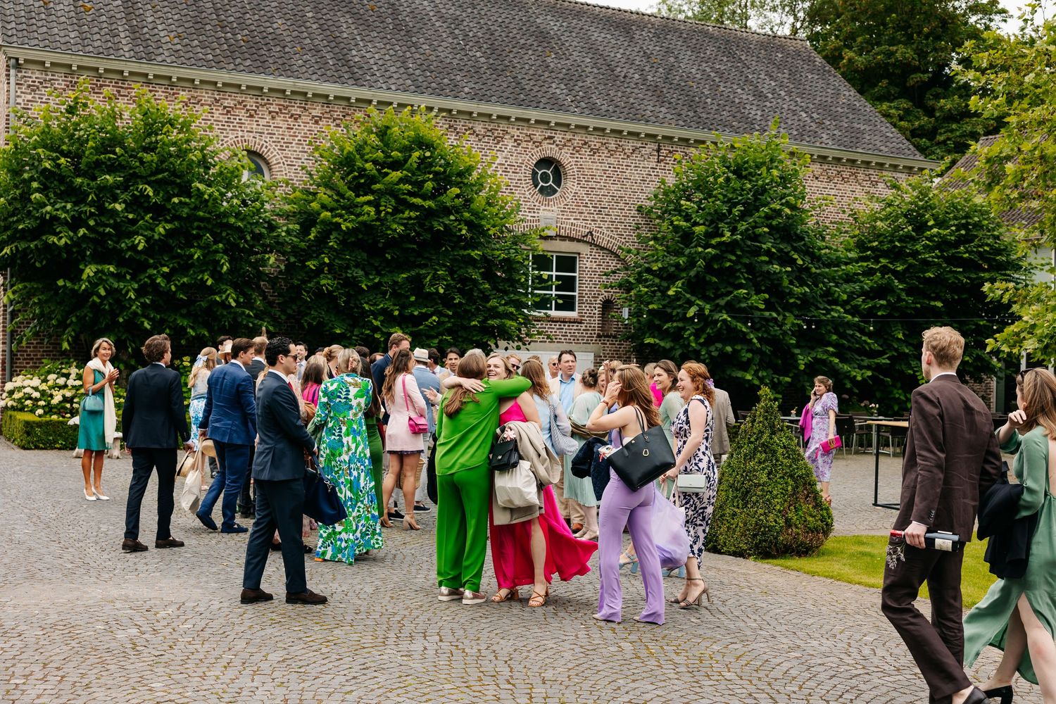 Wedding guests mingling in courtyard outside rustic brick venue surrounded by greenery.