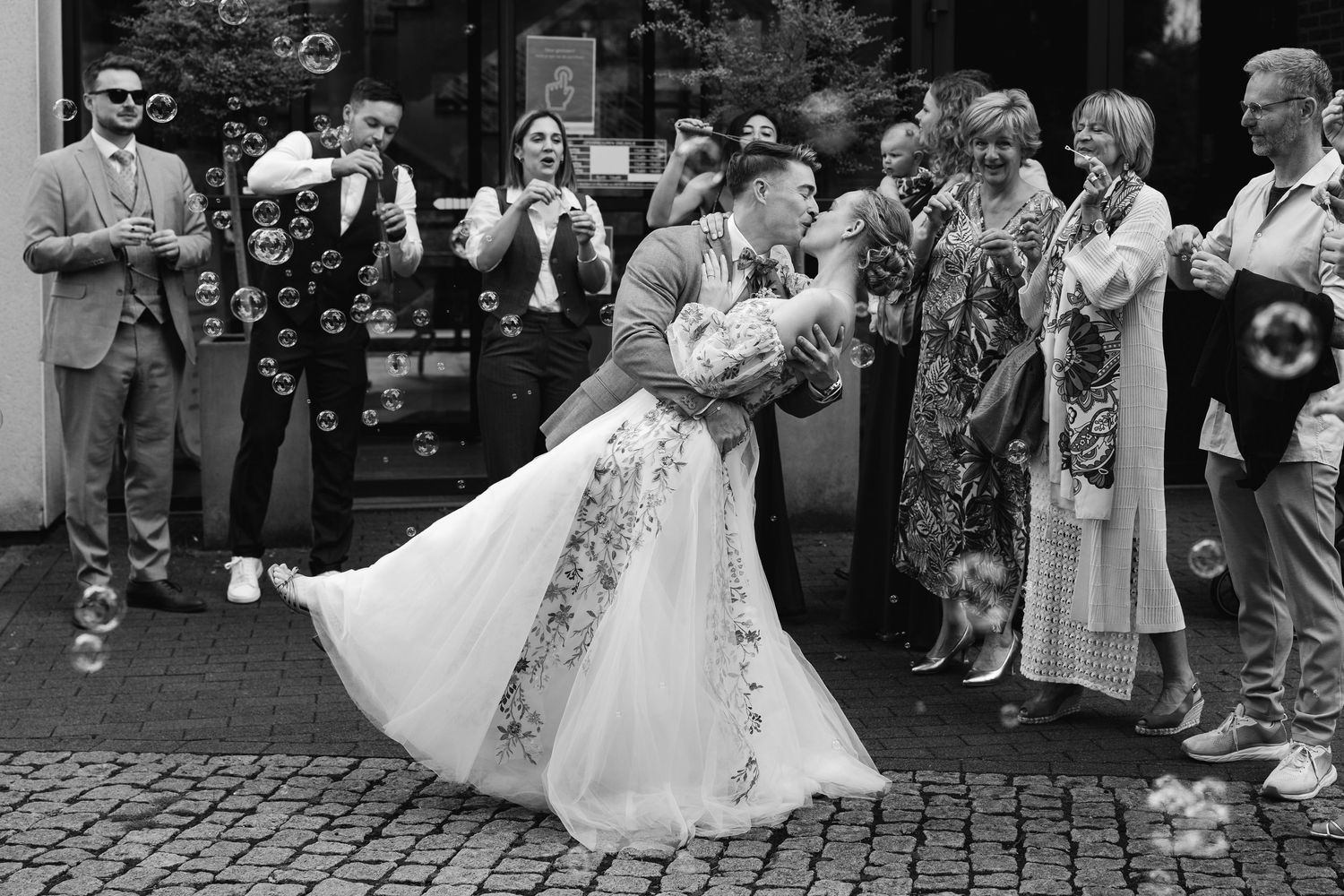 Black and white photo of groom dipping bride in floral dress amid soap bubbles.