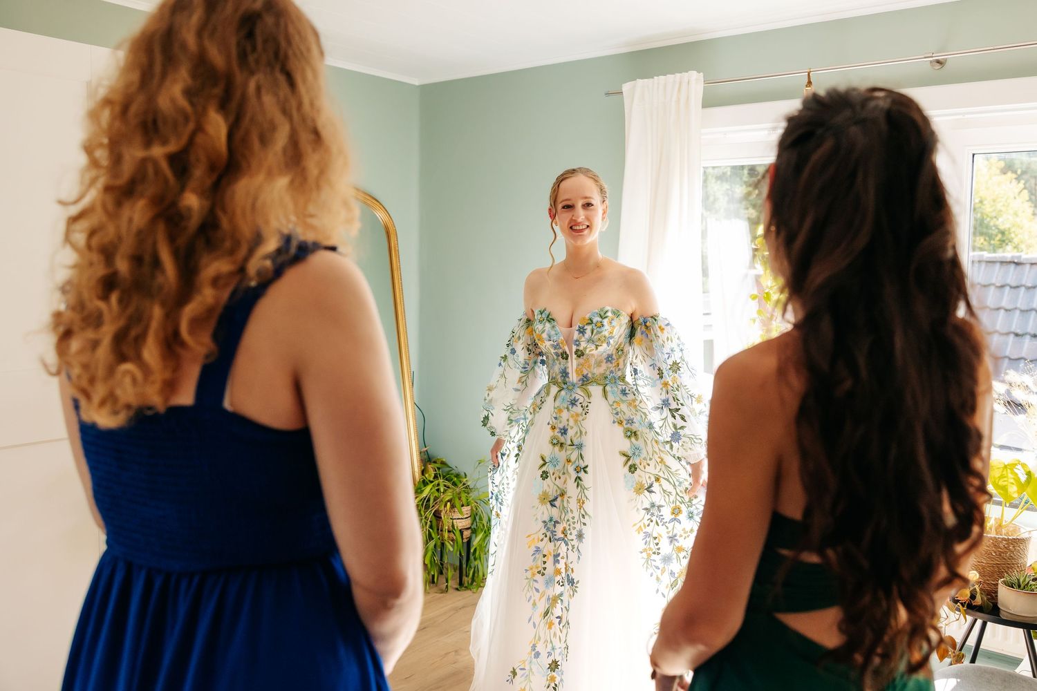 Two bridesmaids watching bride in floral wedding gown stand near sunlit window smiling.