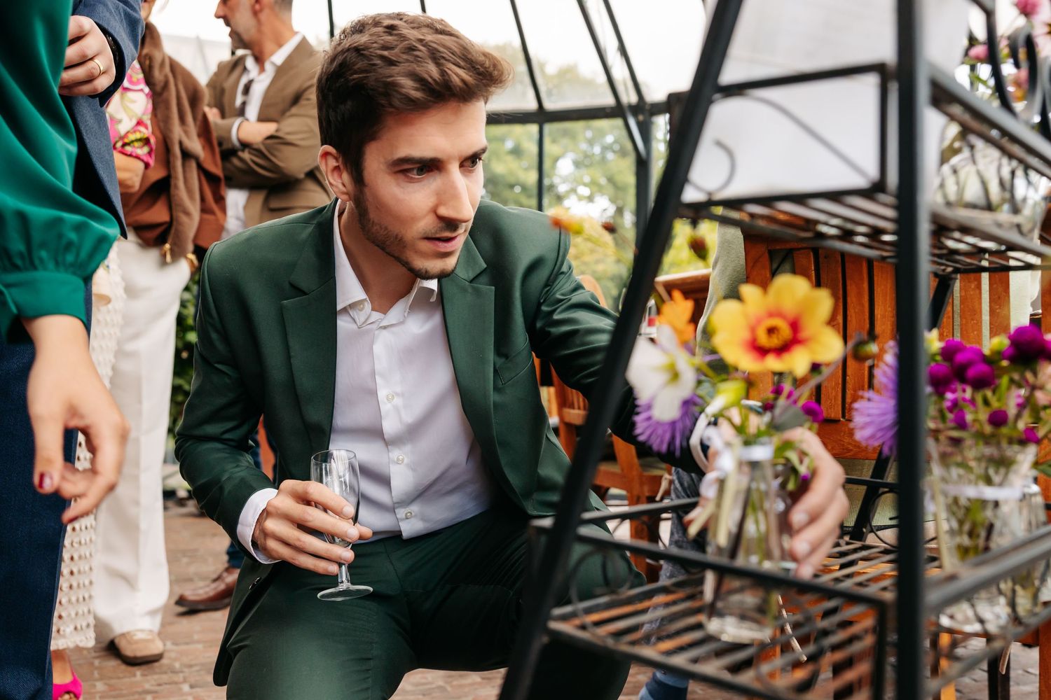 Man in green suit crouching to examine flowers at elegant outdoor wedding reception.