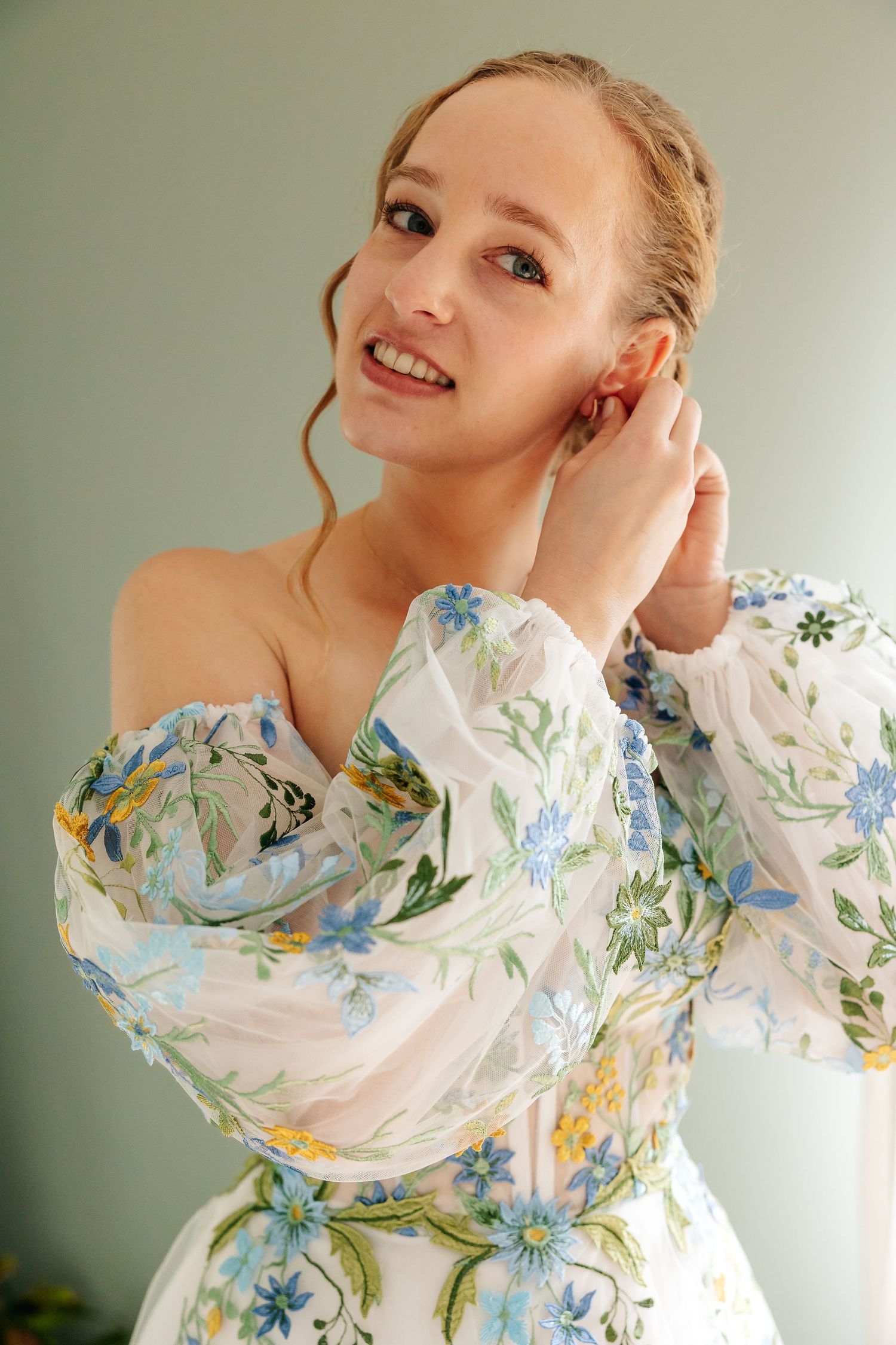 Smiling bride in a floral off-shoulder wedding gown adjusts her earring while getting ready.