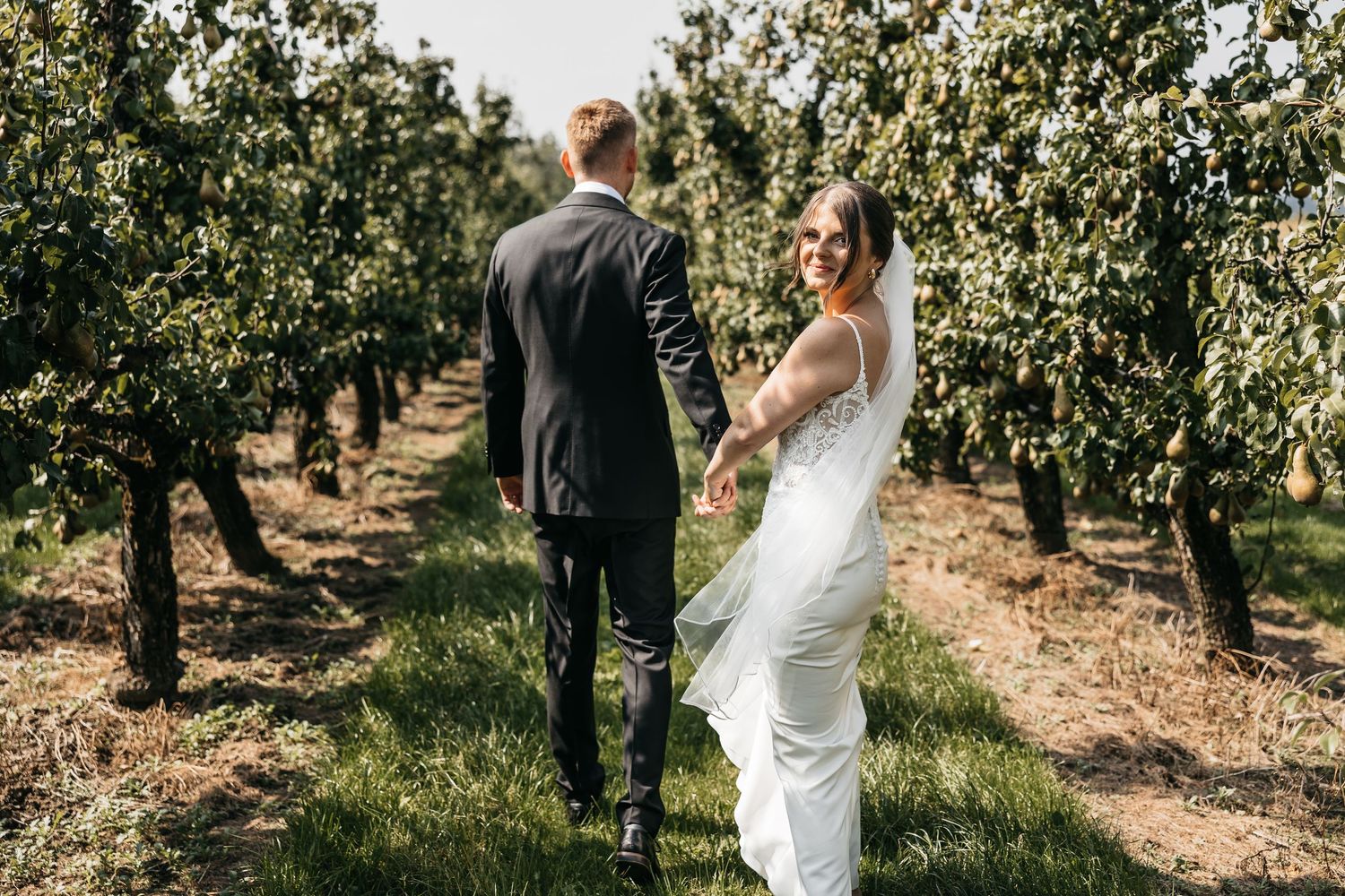 Smiling bride looks back while walking with groom through a beautiful green orchard setting.