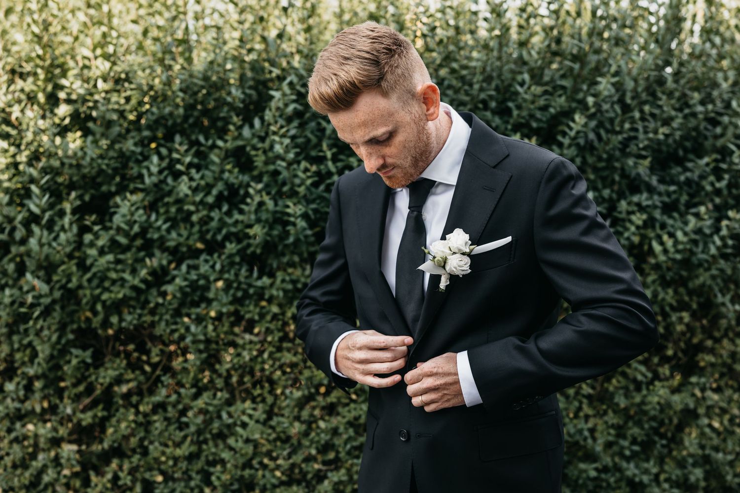 Groom in black suit with white boutonniere adjusting jacket buttons in front of green hedge.