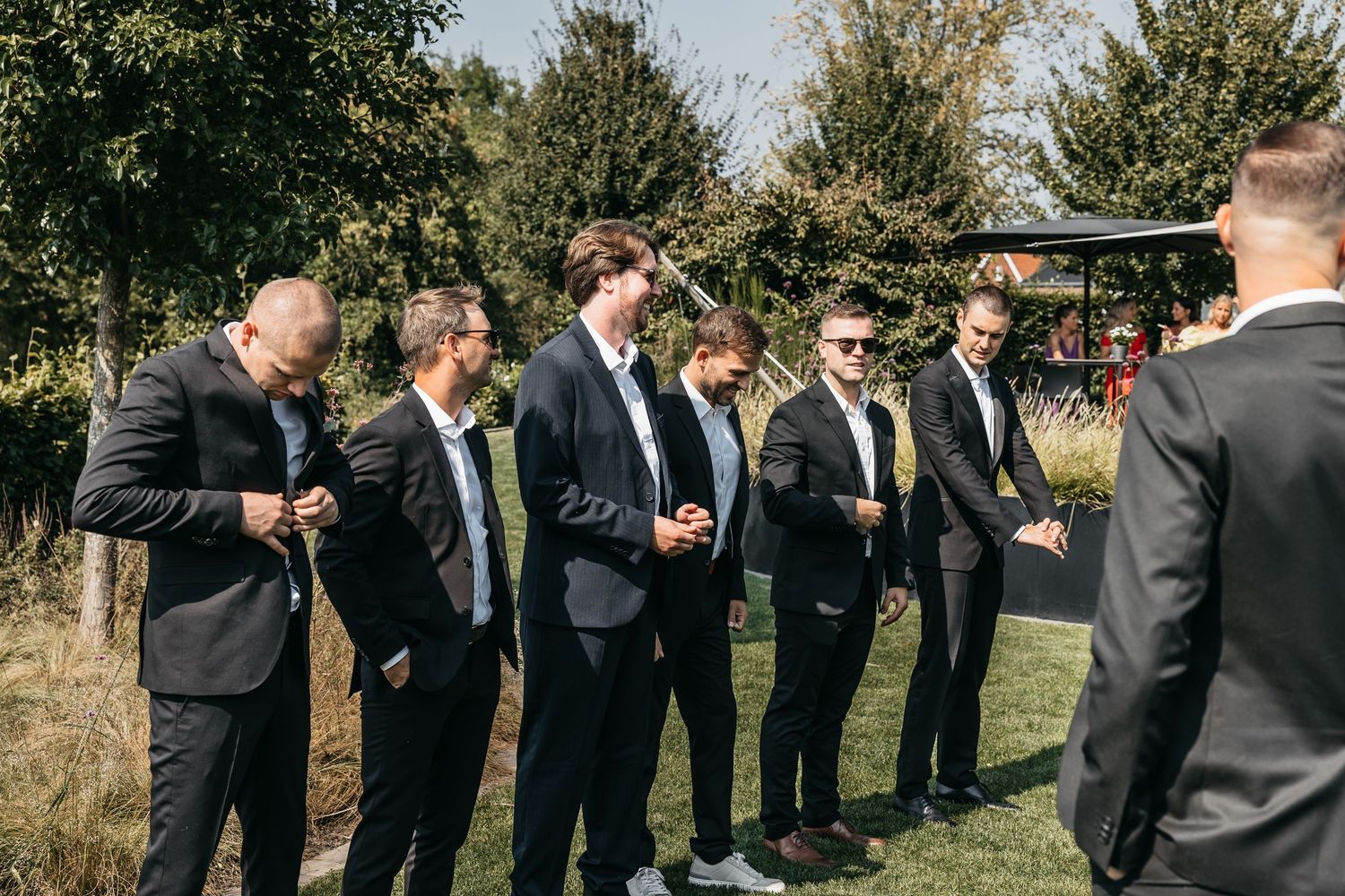 Groomsmen in dark suits adjusting ties and chatting outdoors at a sunny wedding venue.