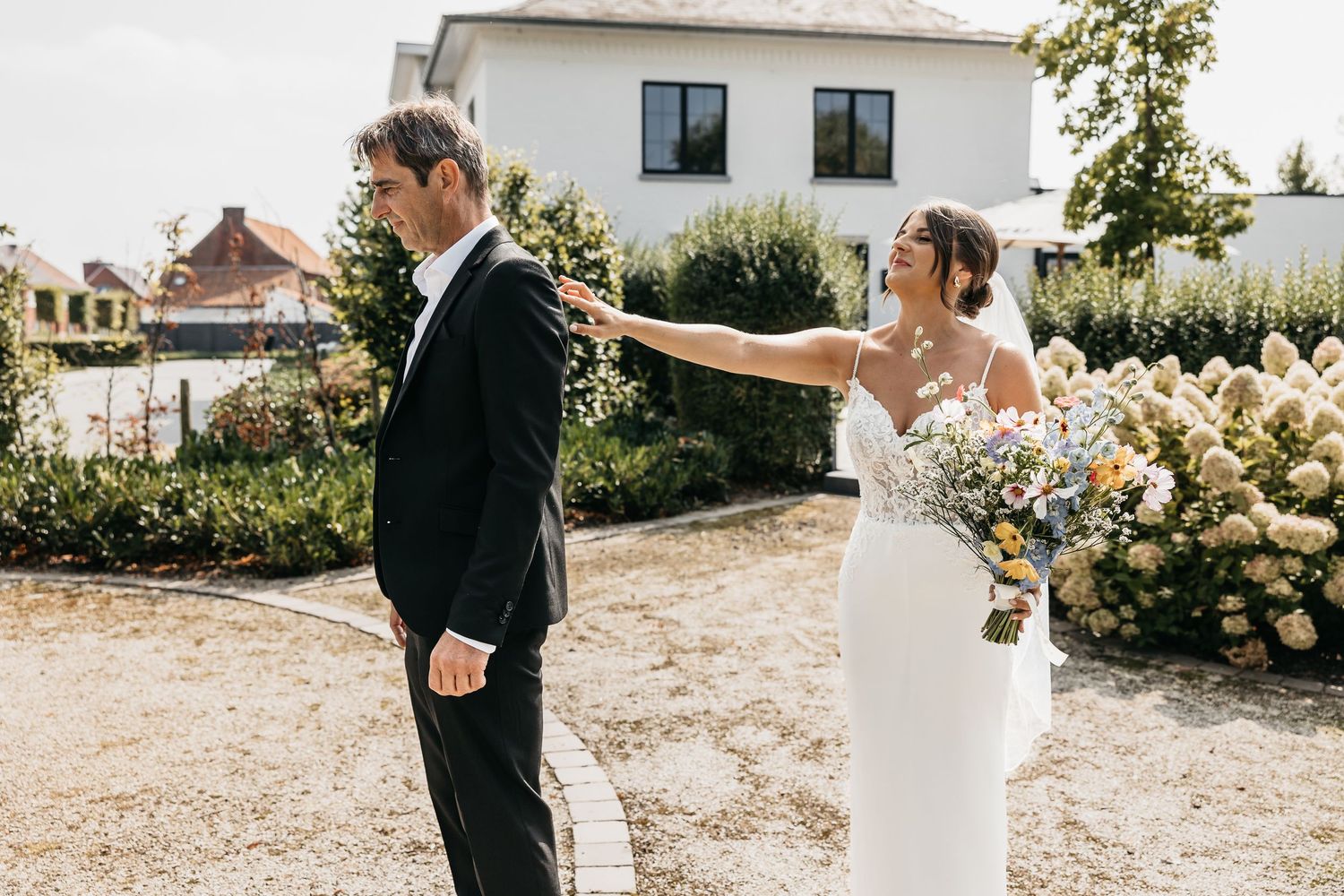 Bride in white gown taps groom's shoulder from behind during first look moment in sunny garden.