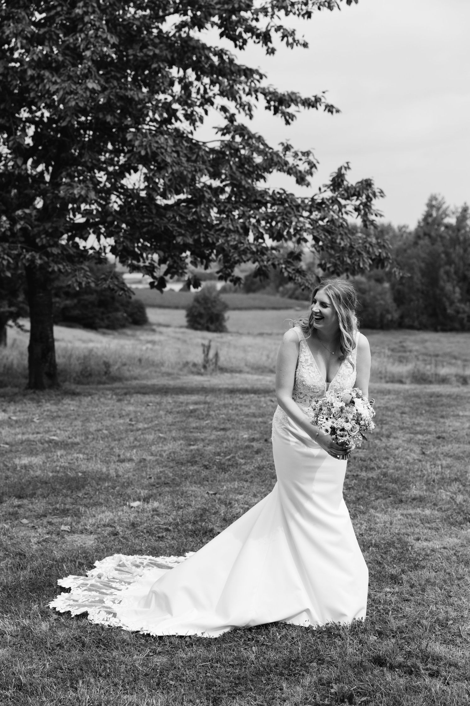 Black and white photo of a laughing bride in a lace gown with a long train outdoors.