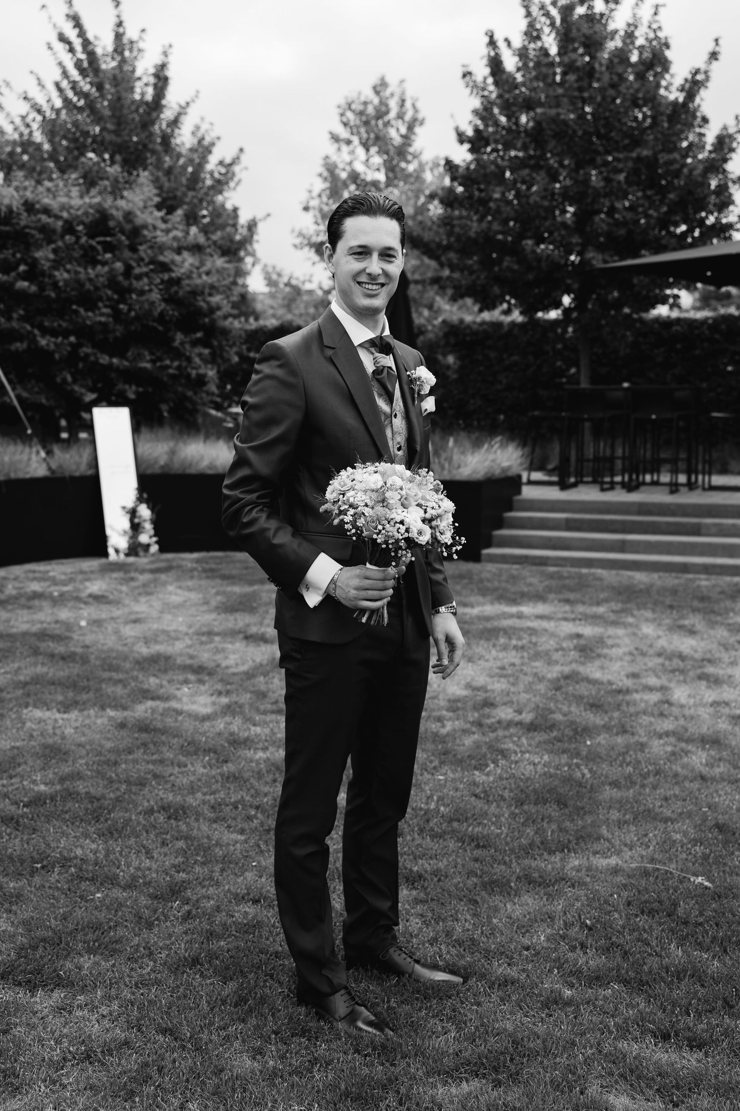 Groom in dark suit holding bridal bouquet smiling outdoors in black and white wedding photo.