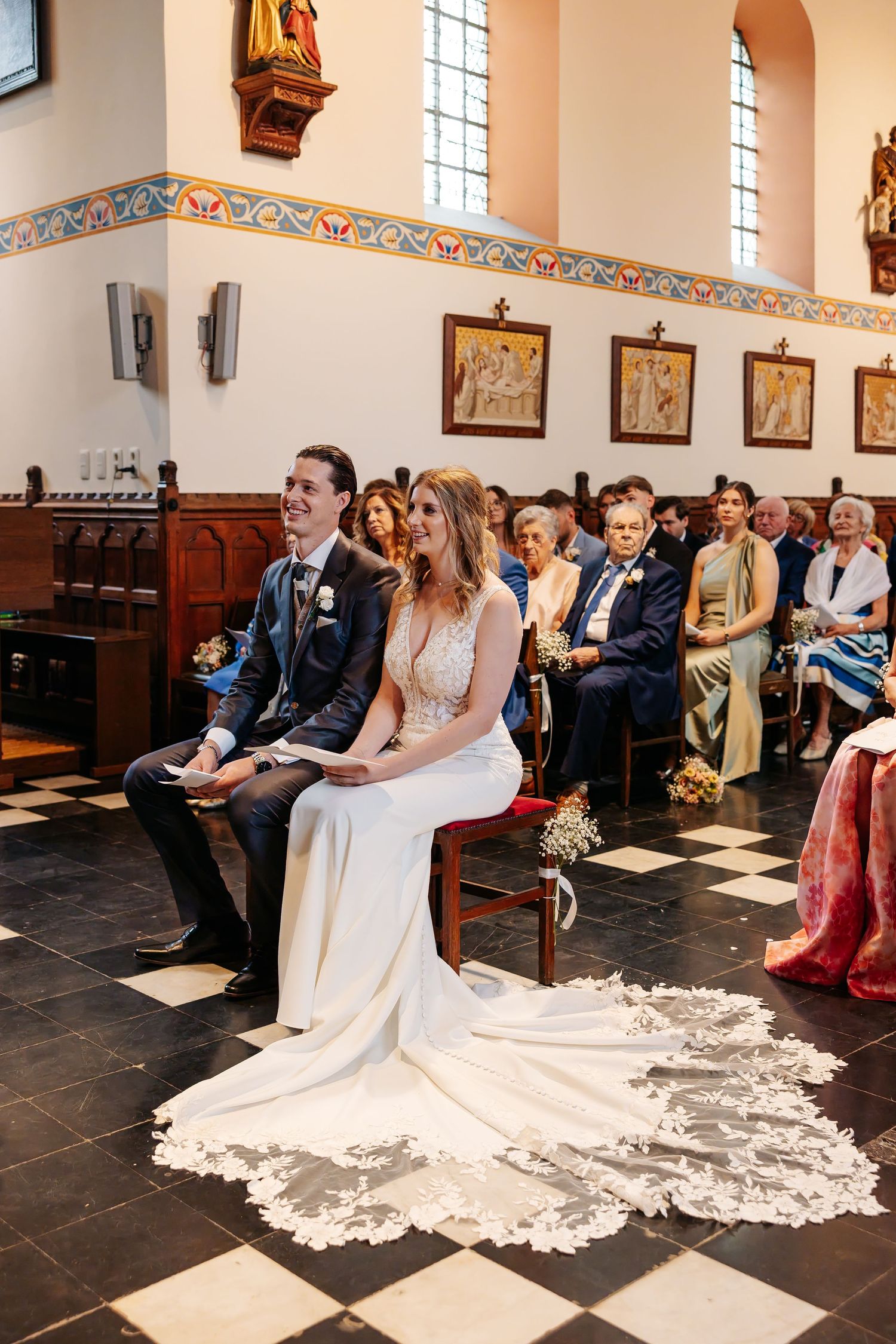 Bride and groom seated at altar during church ceremony with lace train displayed.