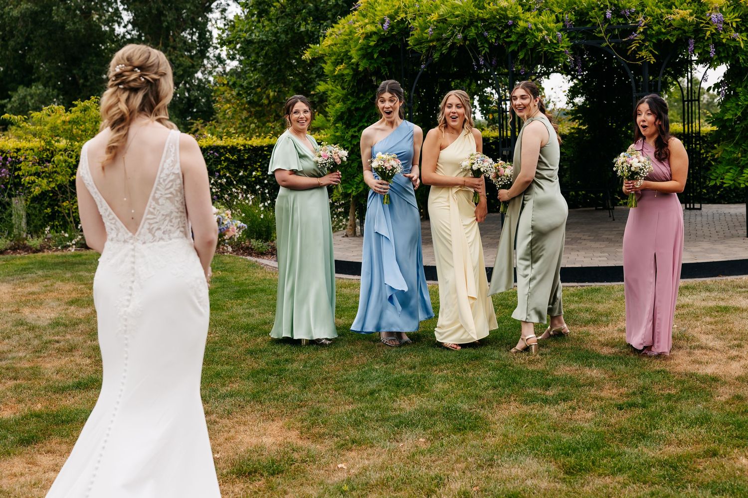 Bride in lace gown faces laughing bridesmaids in colorful pastel dresses on garden lawn.