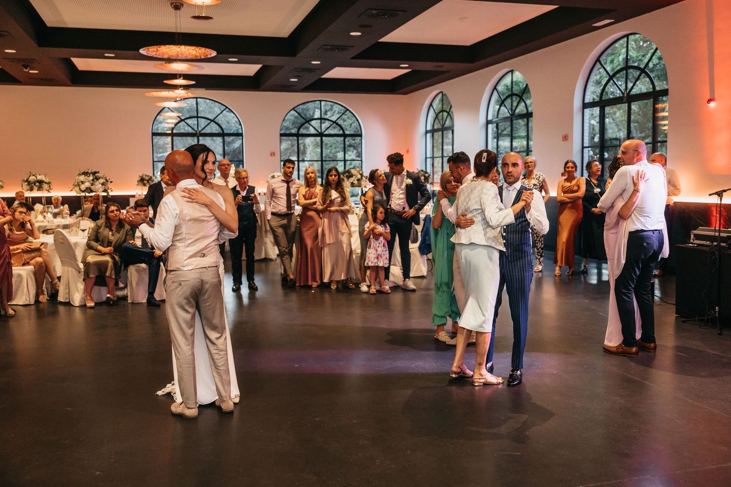Wedding guests dancing together in elegant venue with arched windows during reception celebration.