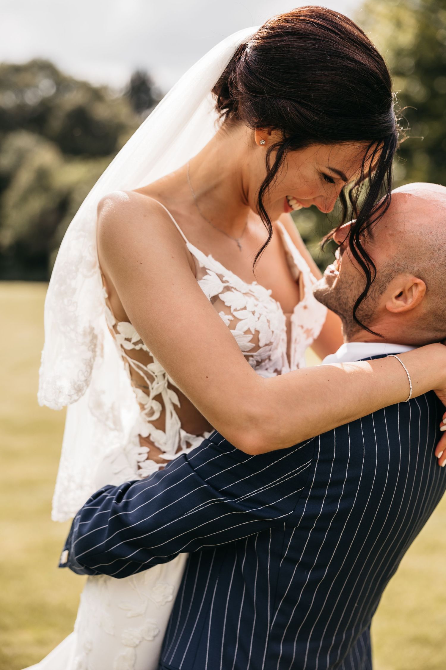 Bride in lace gown and veil smiling while embracing bald groom in navy pinstripe suit.