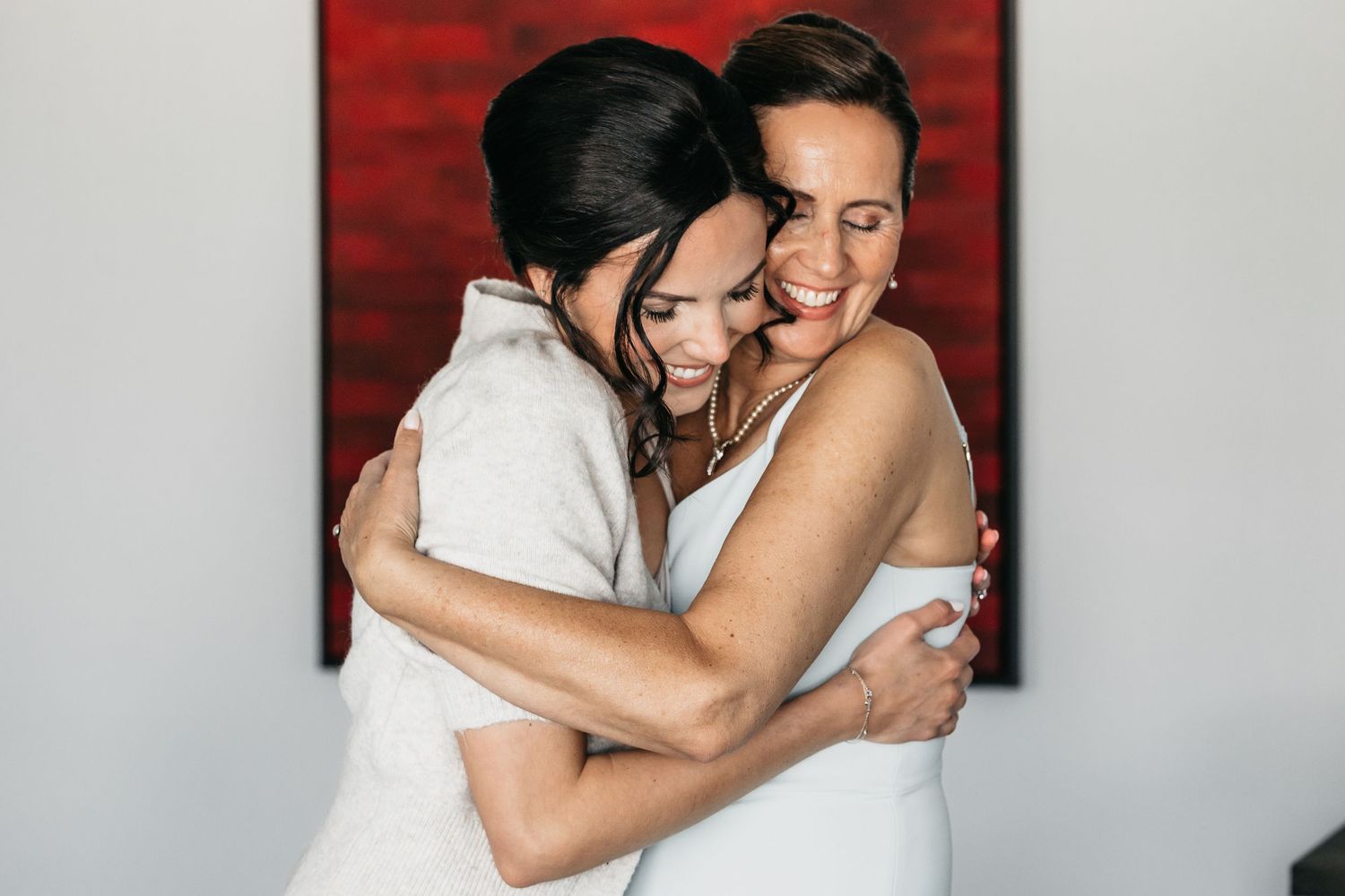 Two women sharing a warm joyful embrace during bridal wedding preparations indoors.