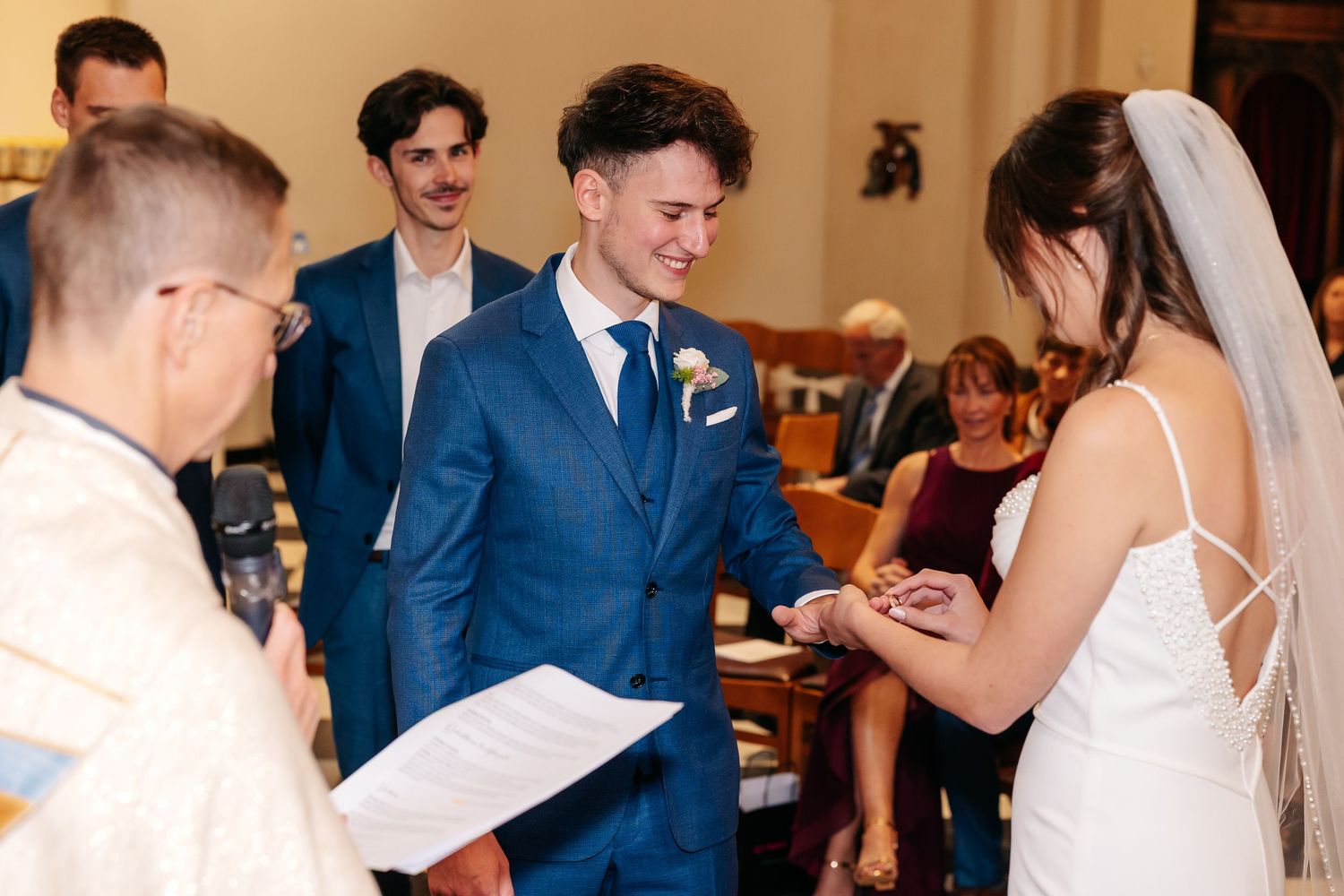 Laughing groom in blue suit receives wedding ring from bride during church ceremony.