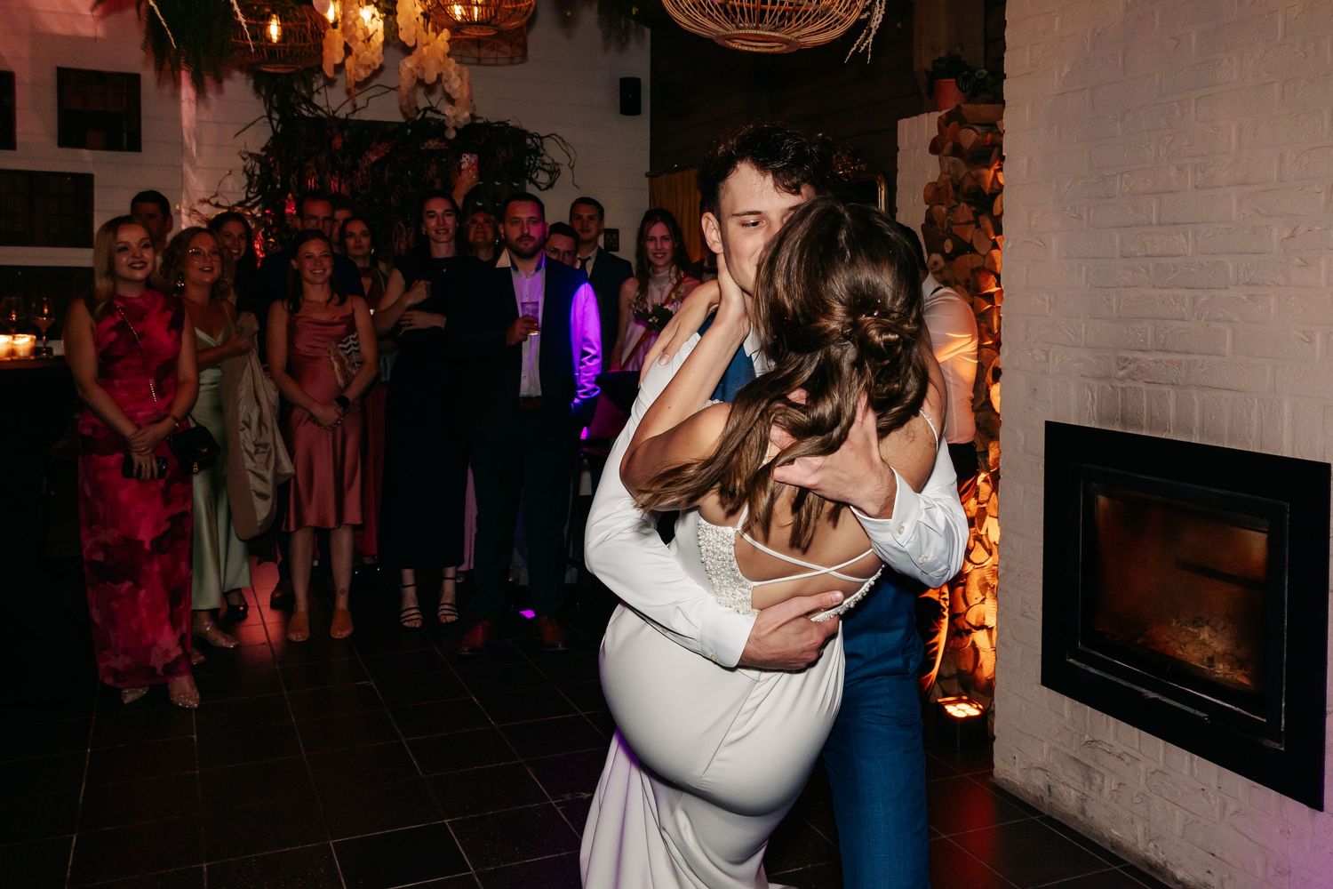 Groom lifts and embraces bride during romantic first dance near fireplace with wedding guests watching closely.