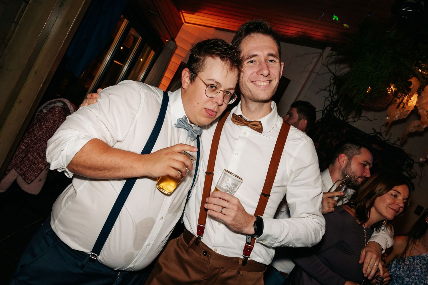 Two men in suspenders and bow ties smile at wedding reception.