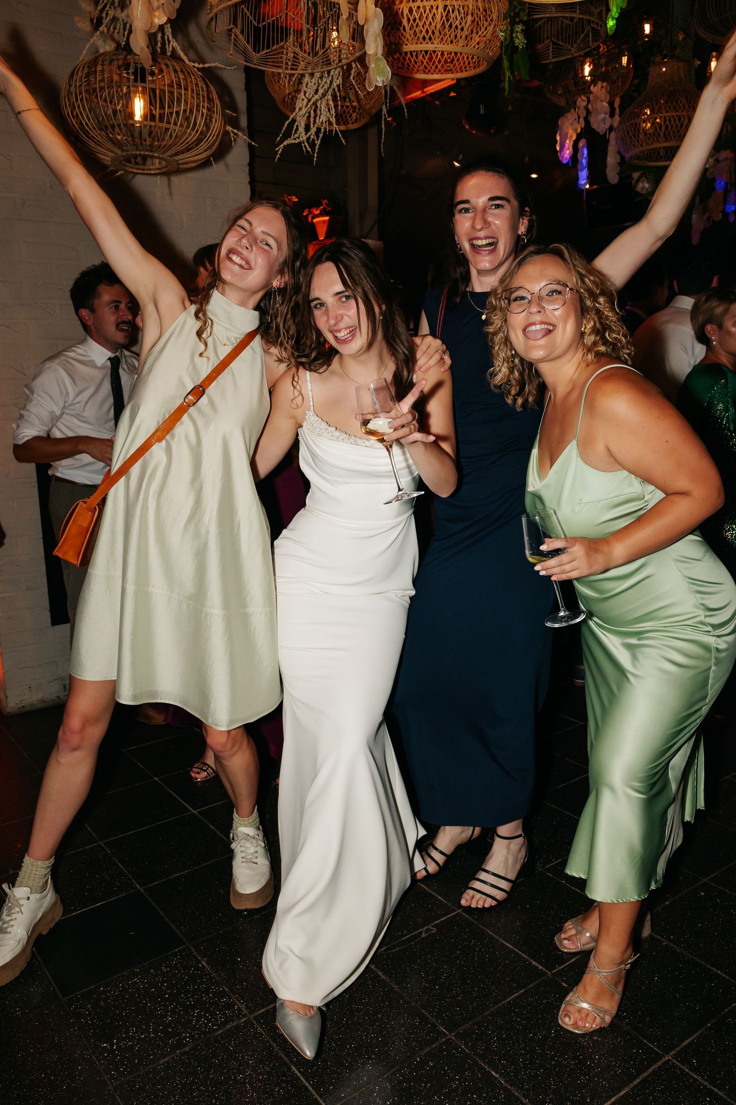 Bride in white gown celebrating with three friends on wedding reception dance floor holding champagne.