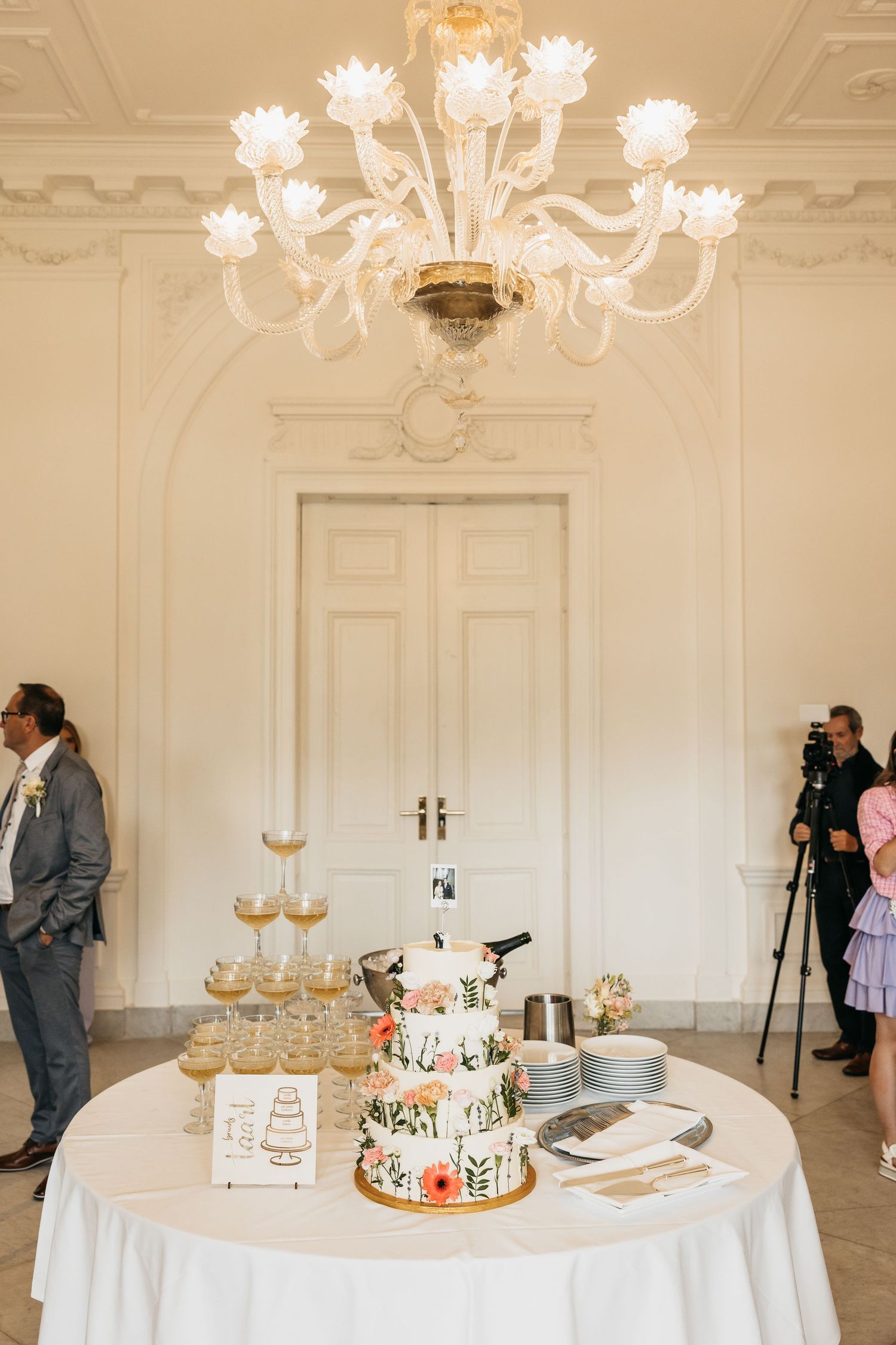 Elegant wedding reception room with chandelier, floral cake, champagne tower on white tablecloth.
