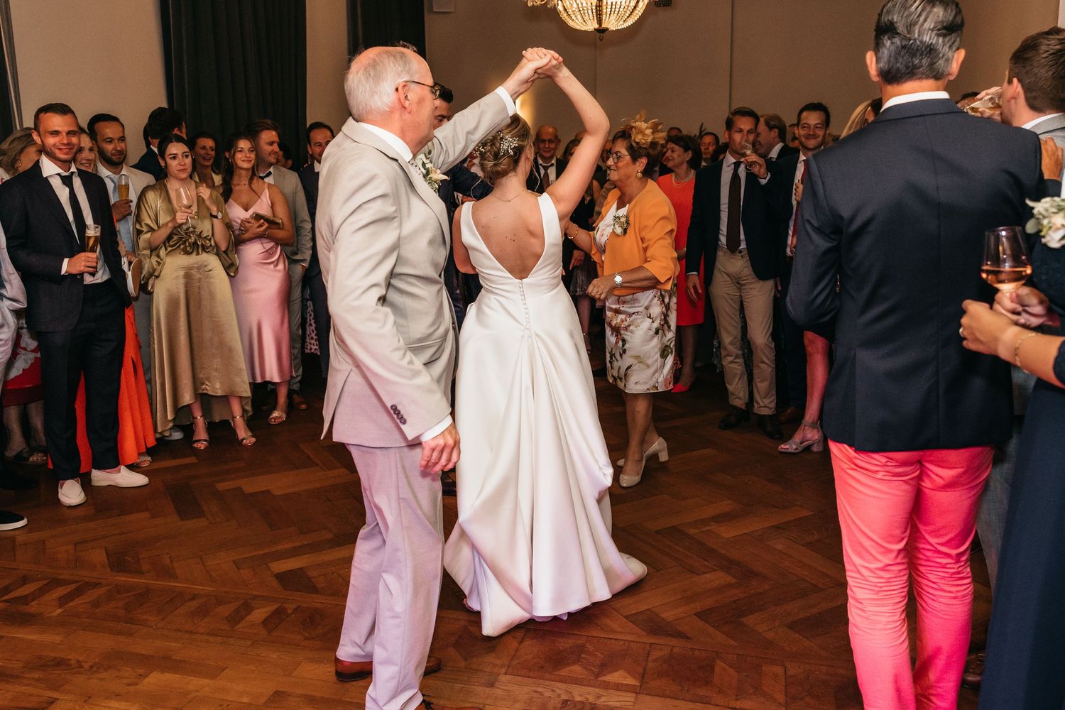 Bride dances with older man in grey suit while guests watch and celebrate at wedding reception.