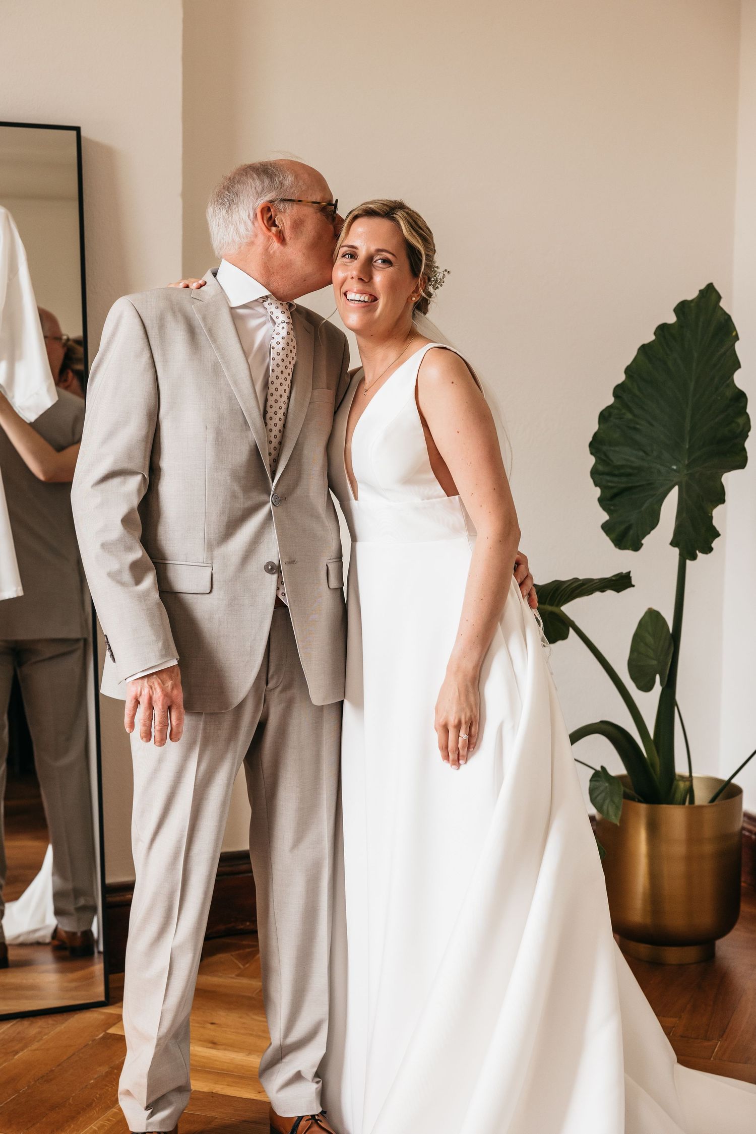 Older man in gray suit kissing smiling bride in white wedding gown on the cheek indoors.