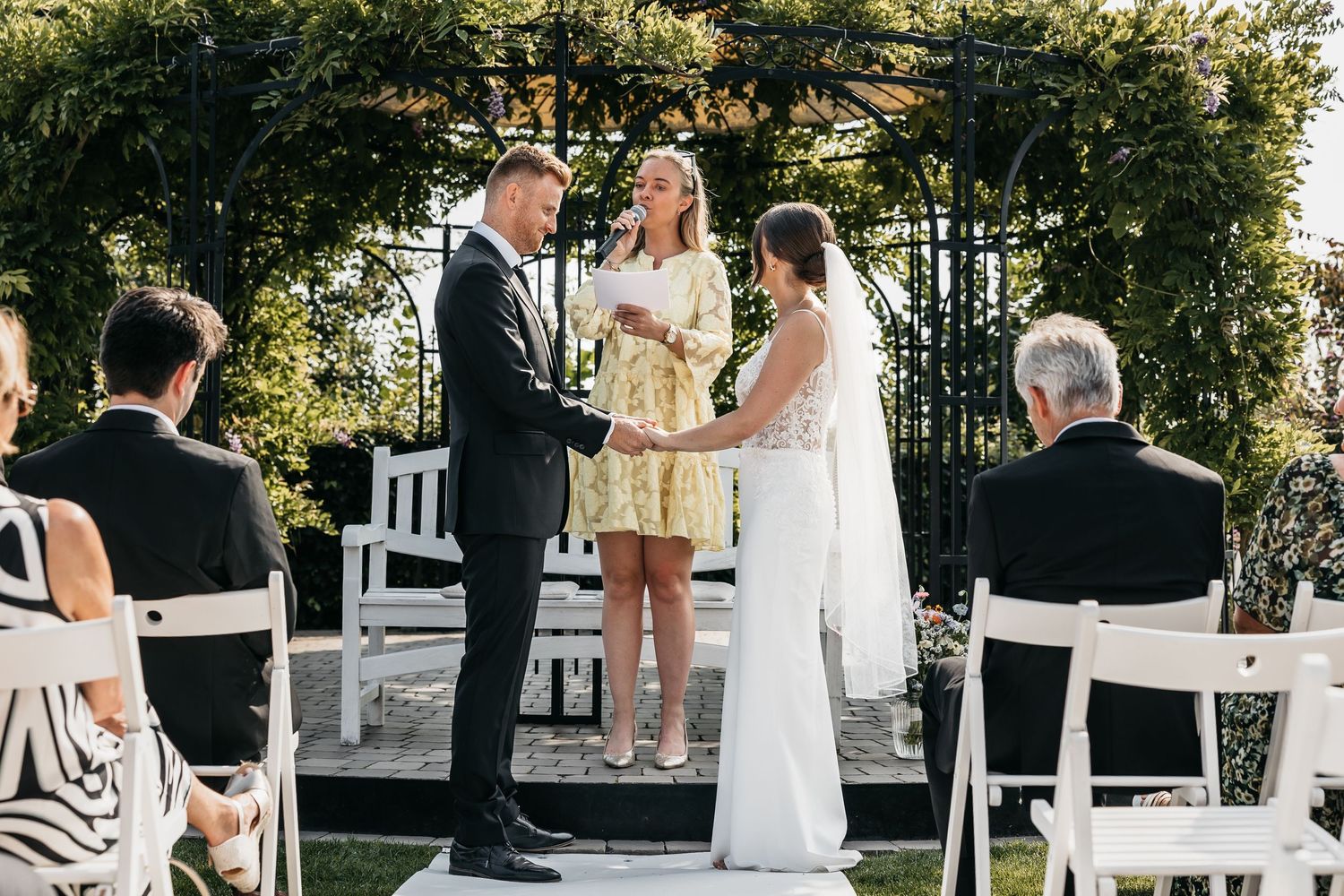 Bride and groom stand at altar with officiant under garden arbor at outdoor wedding.