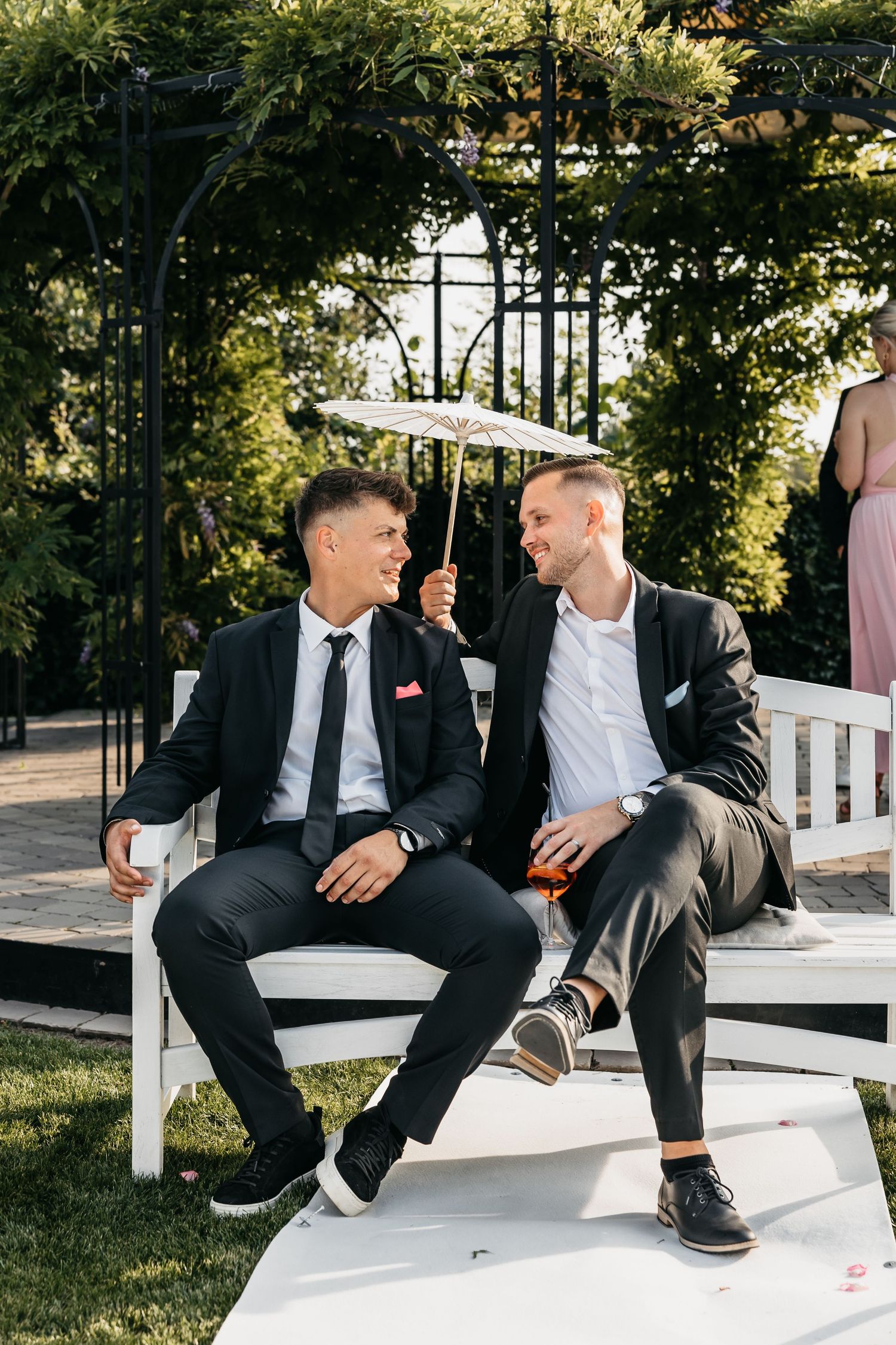 Two smiling male wedding guests share a laugh on a white bench under a decorative umbrella.