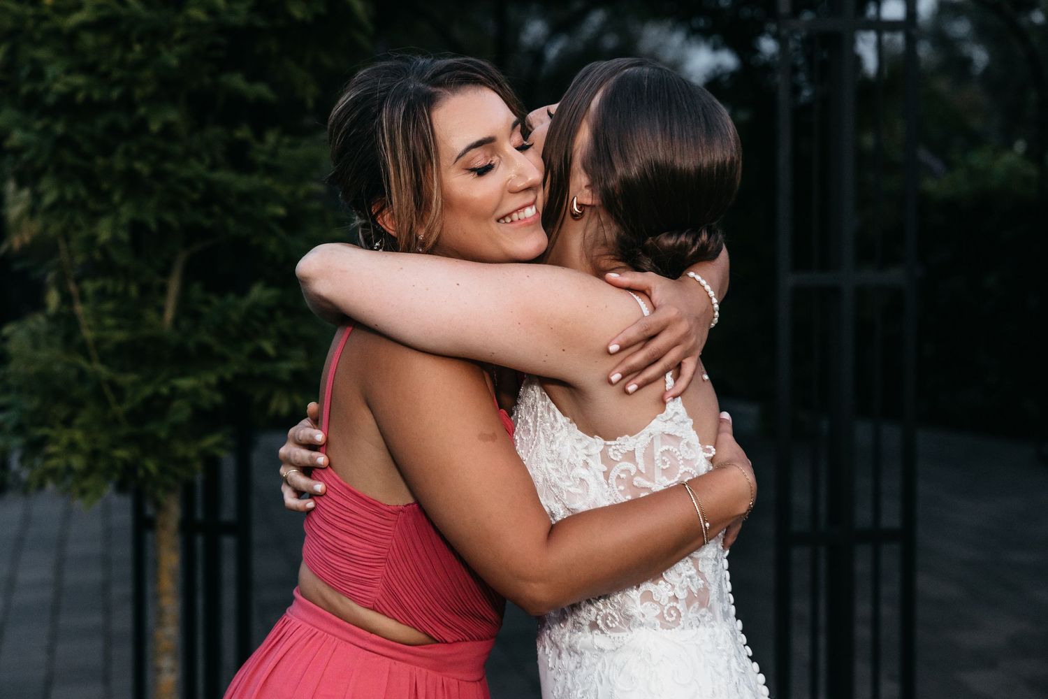 Bridesmaid in pink dress sharing warm embrace with bride in lace wedding gown outdoors.