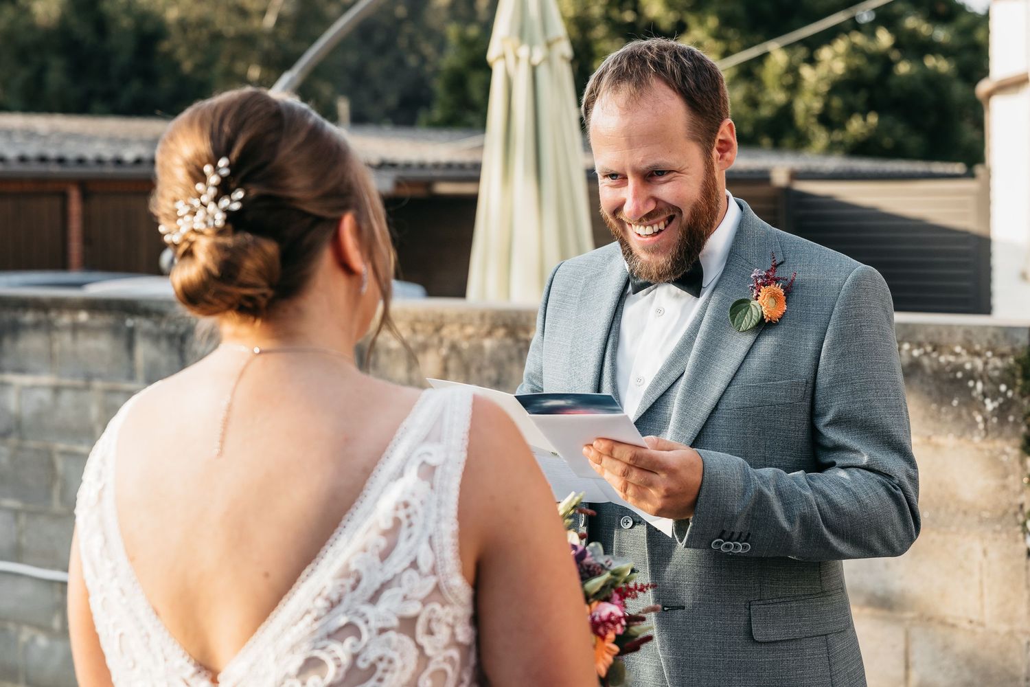 Smiling groom in gray suit reads wedding vows to bride during outdoor ceremony.