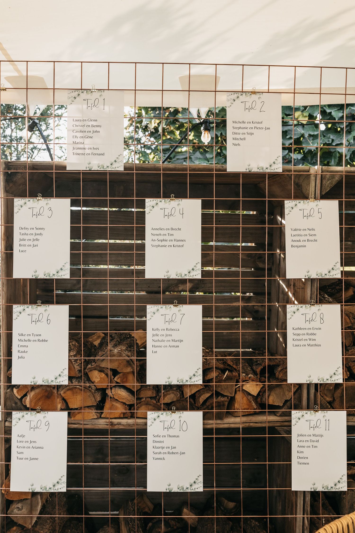 Wedding seating chart cards displayed on rustic metal grid with greenery decorations.