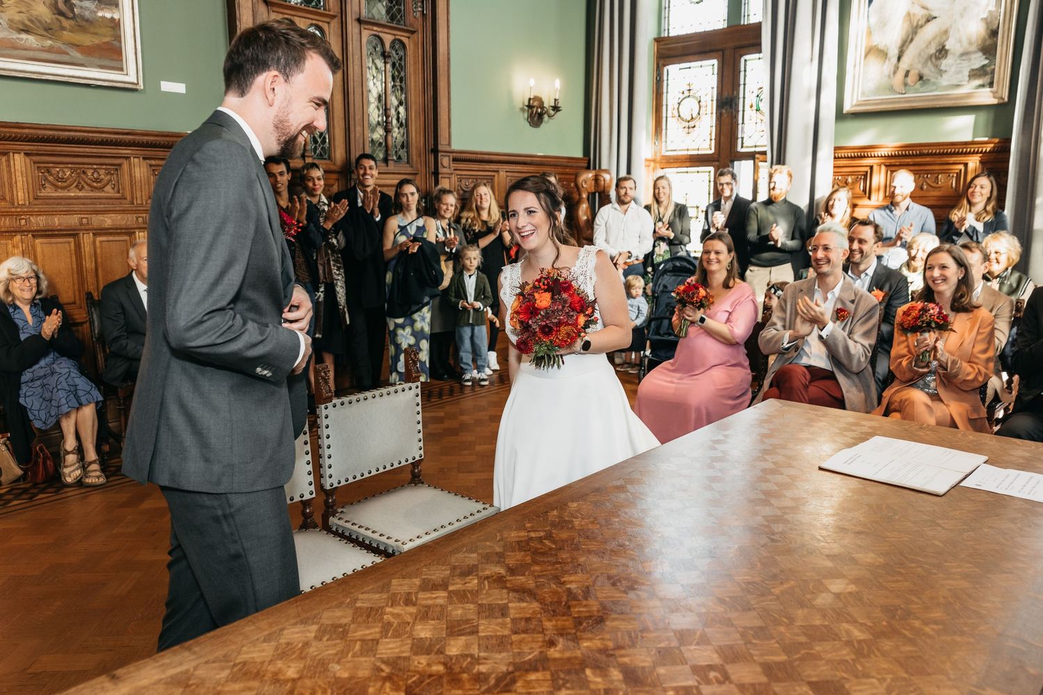 Bride holding red bouquet laughing with groom at a wooden table during civil wedding ceremony with guests.