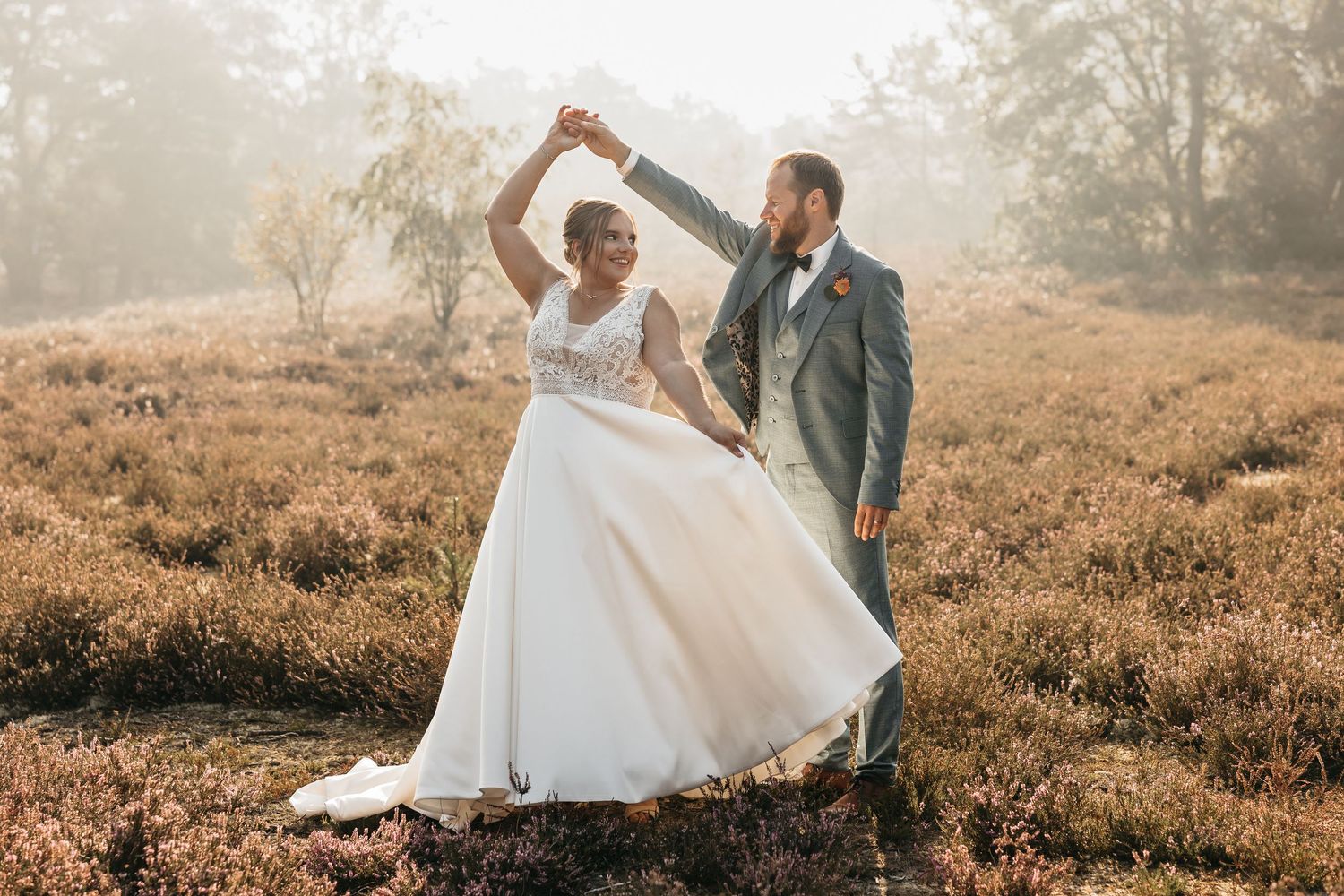 Joyful bride and groom dancing together in golden heather field during misty outdoor wedding shoot.