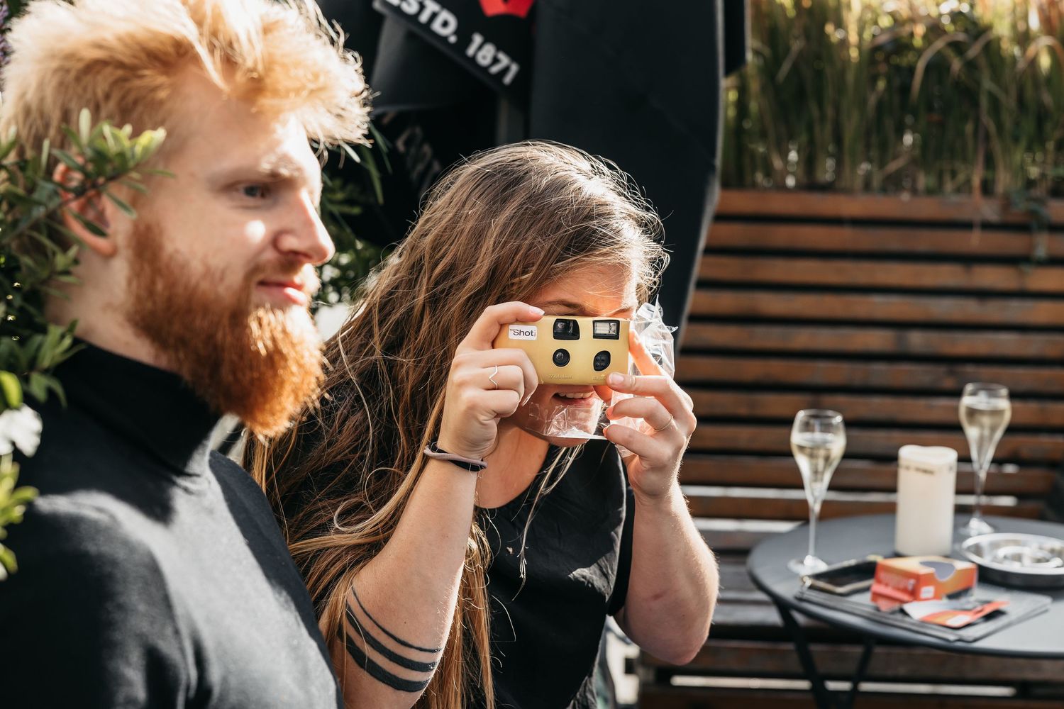 Red-haired bearded man smiling as woman photographs with disposable camera at outdoor wedding party.