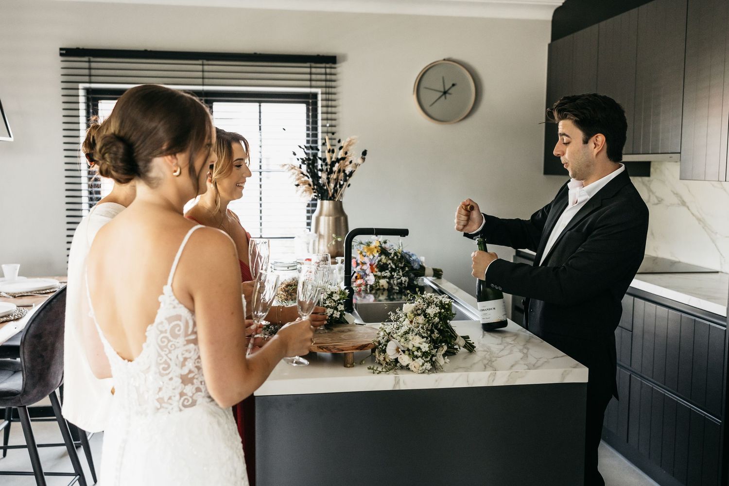 Bride and bridesmaids gathering around kitchen island as groomsman prepares champagne.