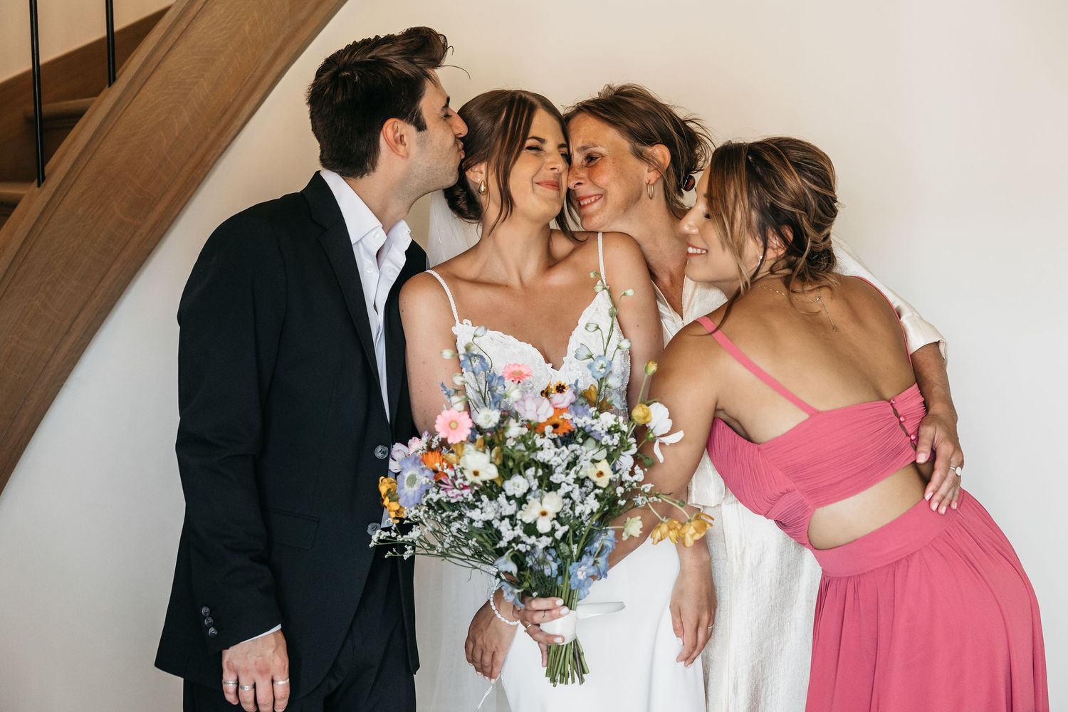 Bride with wildflower bouquet receives kisses from three joyful family members near staircase.