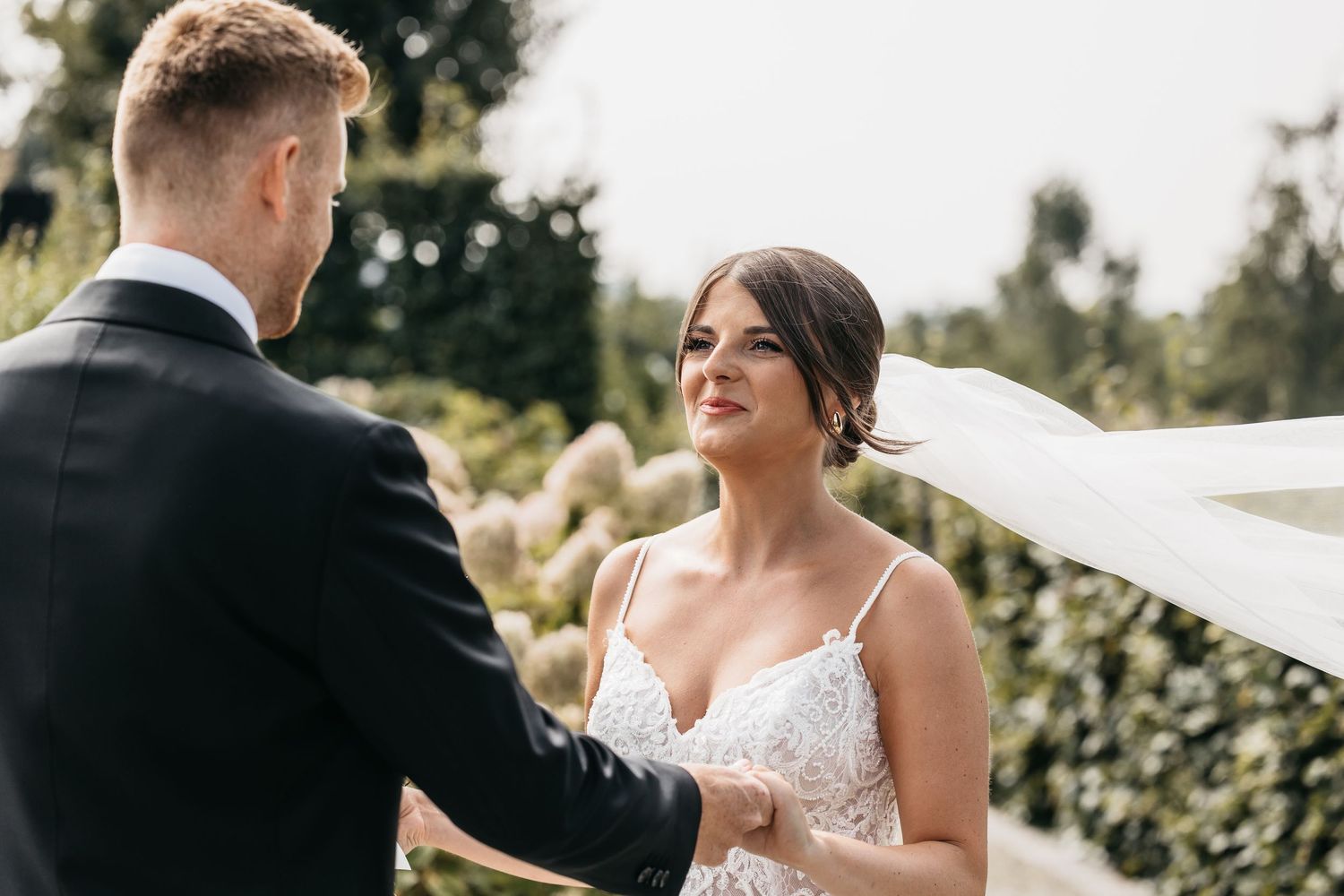 Bride in lace wedding dress smiles lovingly at groom during outdoor wedding ceremony with greenery backdrop.