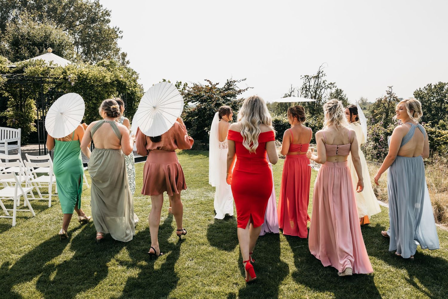 Bridesmaids in colorful dresses holding white parasols walking across garden lawn at sunny wedding.