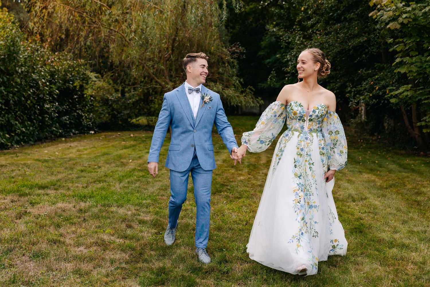 Newlyweds walk hand-in-hand smiling through a lush garden, bride in floral gown and groom in blue suit.