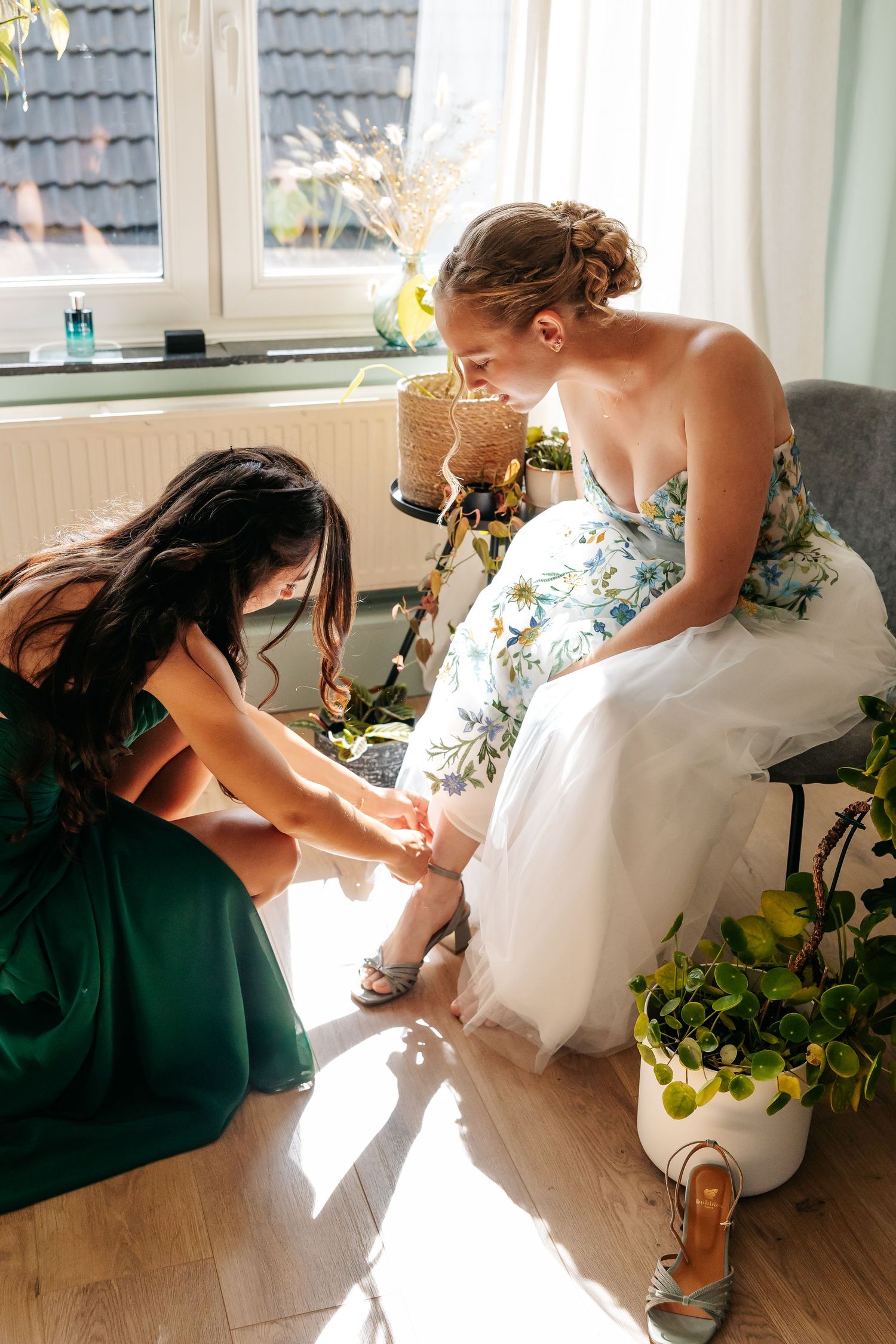 Bridesmaid in green dress helping bride fasten strappy heels while seated in sunlit room.