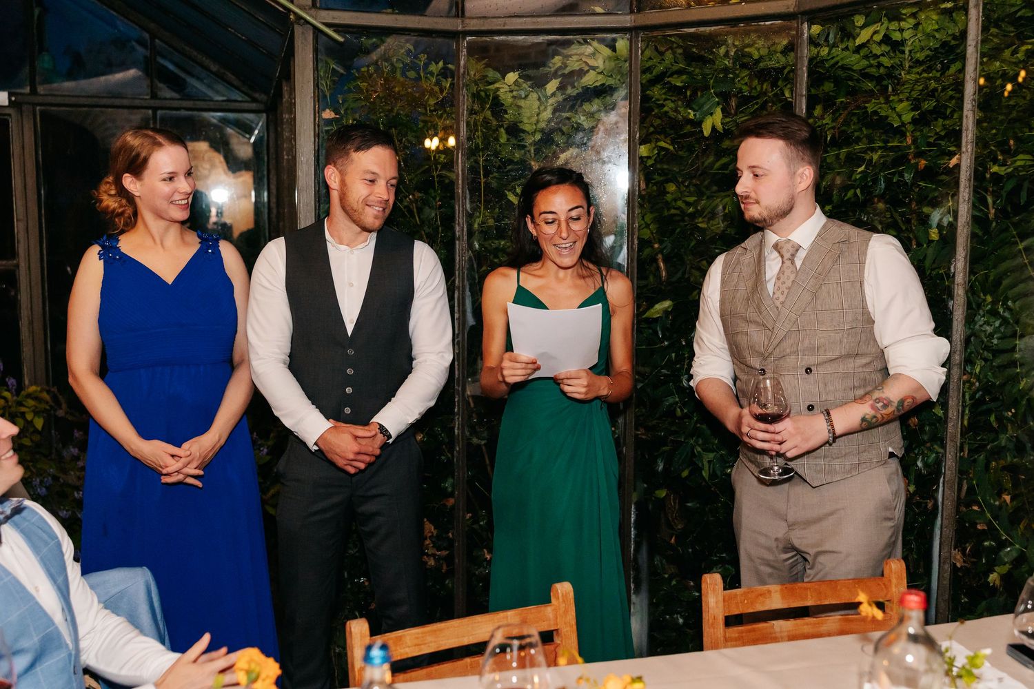 Four wedding guests stand together during a reception speech, including a woman in blue and a man in a grey vest.