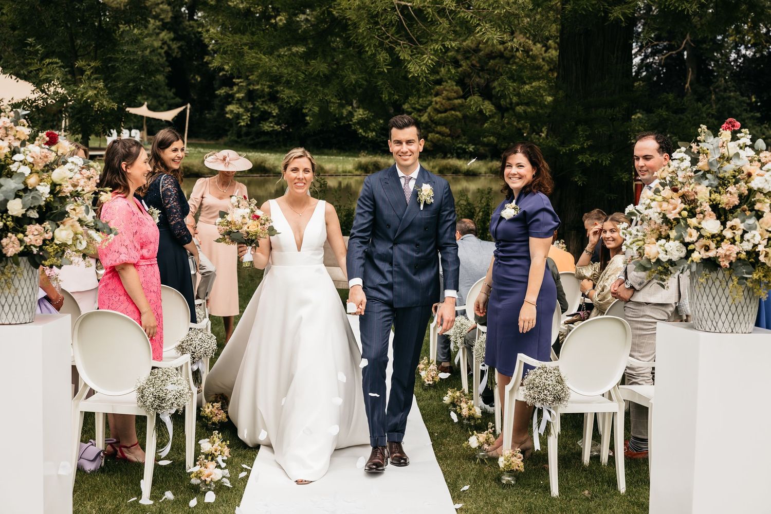 Newlyweds walk joyfully down outdoor aisle together after wedding ceremony surrounded by floral arrangements and guests.