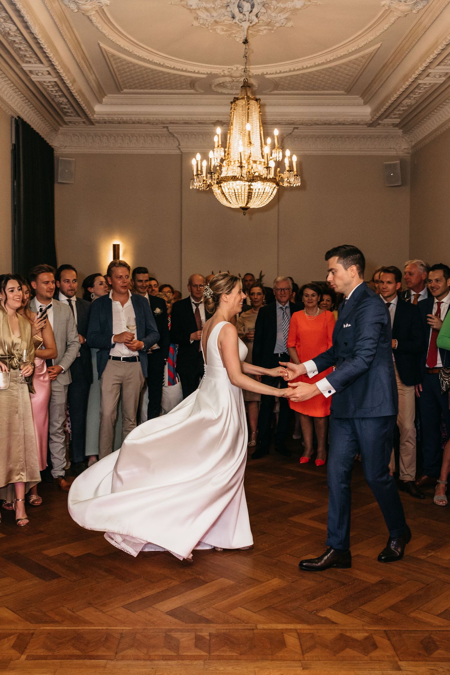 Bride and groom share first dance under crystal chandelier surrounded by cheering wedding guests.