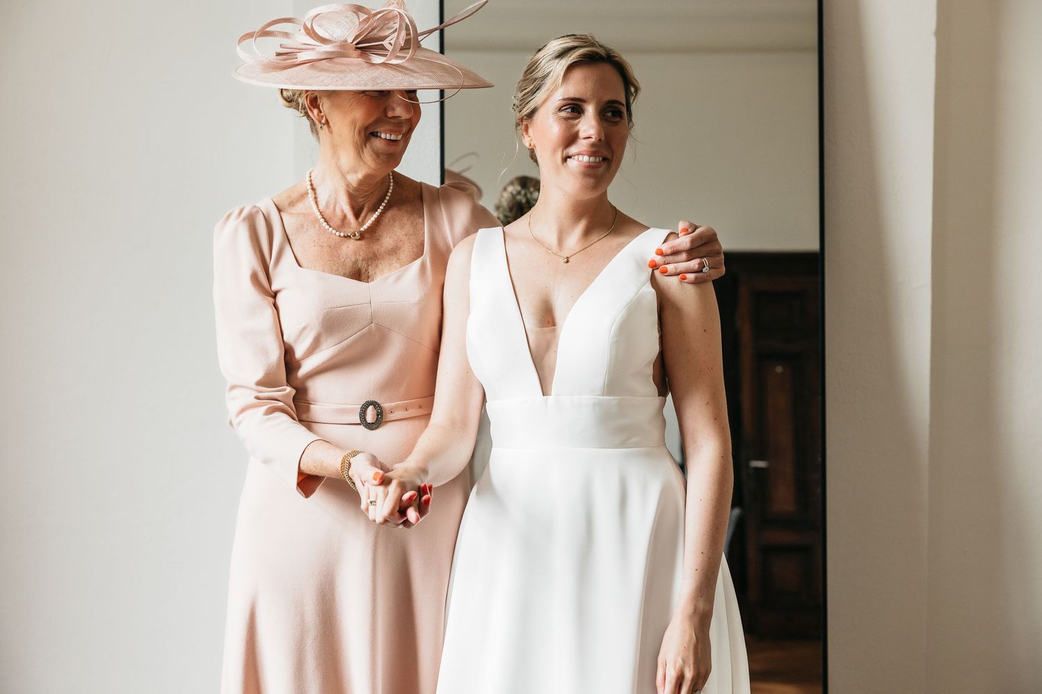 Bride in white gown with older woman in pink dress and fascinator hat smiling together.
