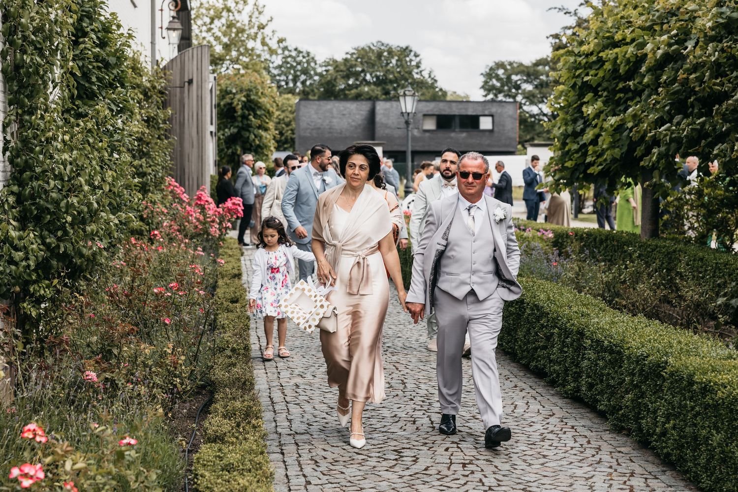 Wedding guests walk along cobblestone garden path lined with roses and manicured hedges.