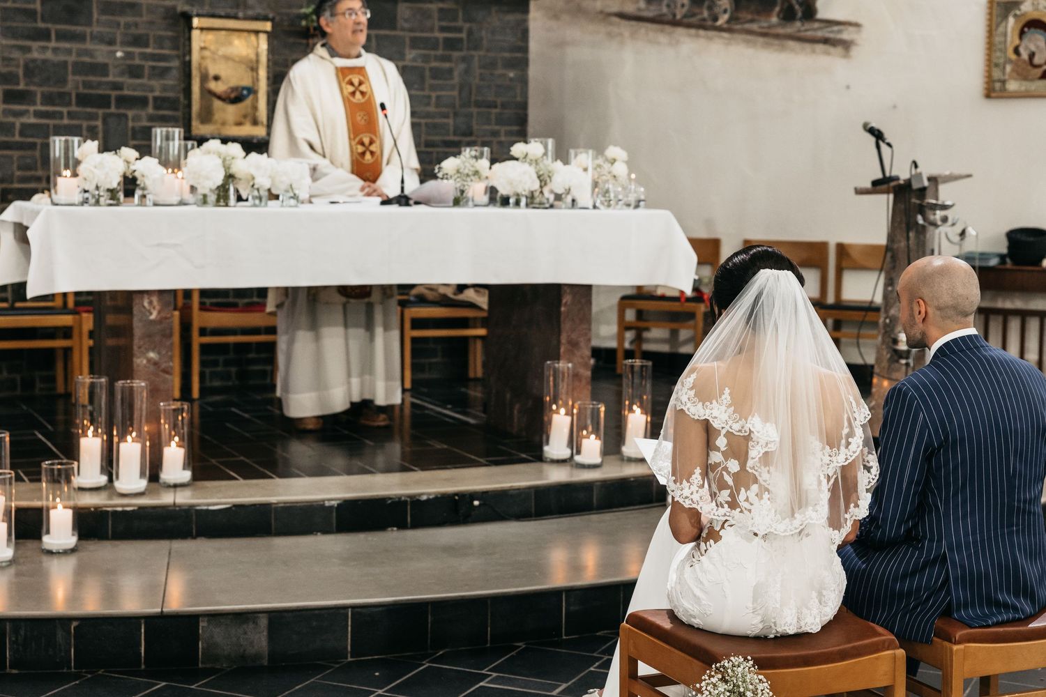 Bride and groom kneel before priest at candlelit church altar during ceremony.